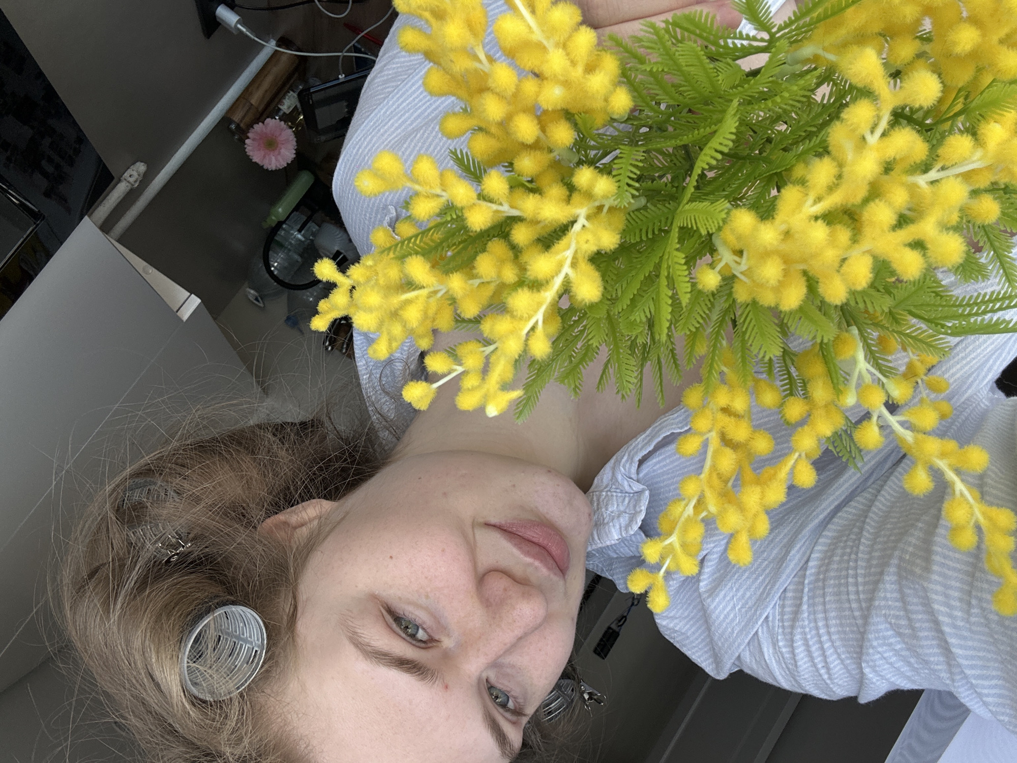 A young Caucasian woman with fair skin and brown hair styled with rollers poses for a selfie, holding a bouquet of yellow mimosa flowers. The background includes a portion of a room with a cabinet, various items on a counter, including a pink flower, and some other objects. The woman is wearing a blue and white striped shirt. The photo was taken indoors and is rotated counter-clockwise. There is no information on the city of Kazan in the image.