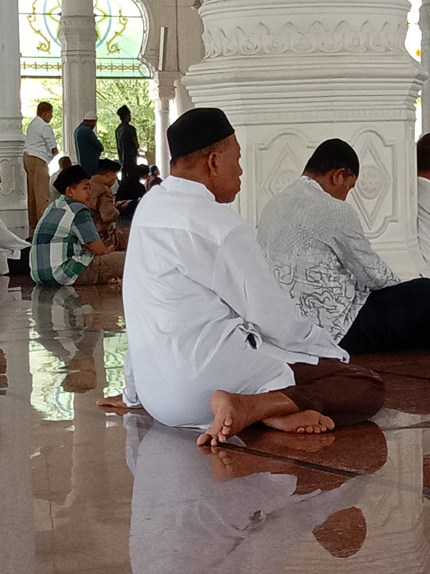 A group of men and a child are sitting on the floor of a building, likely a mosque, in Lhokseumawe, Indonesia. The floor is highly reflective, possibly due to being wet or polished, and it mirrors the figures and architectural elements. In the foreground, a man in a white shirt and black cap is seated with his back to the camera, his feet visible. To his right, another man, also in a white shirt, is partially visible. To the left, a child in a checkered shirt sits cross-legged. Other individuals are visible in the background, some standing and some sitting. The architecture features white pillars and stained-glass windows with intricate designs. The overall scene suggests a place of worship or communal gathering.