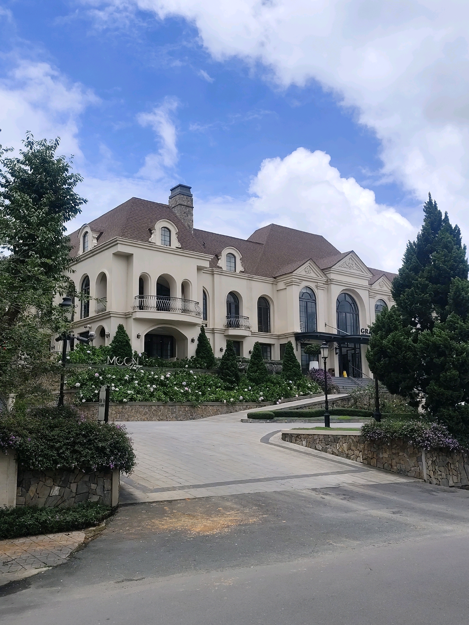 A grand, beige building with a brown shingled roof and arched windows stands prominently. A stone chimney is visible on the left side of the roof. The building is framed by lush green trees and flowering bushes. A sign reading "MOON" is positioned in front of a section of the building with balconies. Another sign near the entrance reads "GRAND". The scene is set against a backdrop of a blue sky with scattered white clouds. The foreground features a paved driveway leading to the building, bordered by stone walls and landscaping.