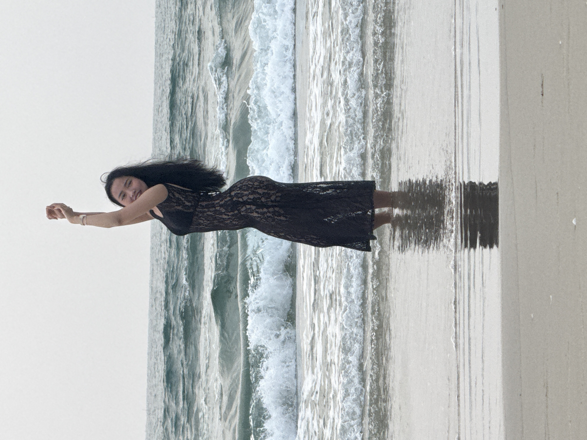 A woman with long, dark hair is seen standing on a beach at Đà Nẵng, Vietnam. She is wearing a black, semi-sheer lace dress. The ocean waves are visible in the background, and her reflection is cast on the wet sand. Her arms are extended outwards, and she is smiling towards the camera. The sky is overcast.