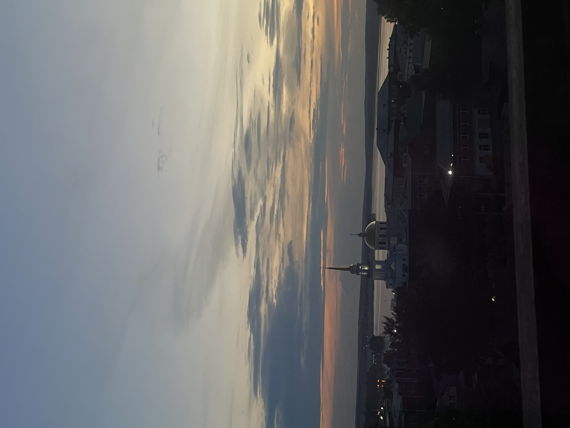 The image captures a sunset over a city, with Votkinsk, Russia as the likely location. The sky is filled with dramatic clouds, painted in hues of pale blue, grey, and orange, indicating either sunrise or sunset. In the middle ground, a distinctive building with a golden dome and a tall, slender spire stands prominently. This structure, likely a religious building such as a church or mosque, is illuminated, suggesting it's either dusk or dawn.
Beyond the domed building, the cityscape unfolds with various structures and trees. The buildings appear to be a mix of residential and possibly commercial, with some showing lights, reinforcing the idea of the day's end or beginning. The dark silhouette of trees frames parts of the scene, adding depth.
In the background, a body of water, likely a river or lake, is visible, reflecting the subtle colors of the sky. The overall atmosphere is serene and picturesque, capturing a quiet moment as day transitions into night. The composition is vertical, with the sky dominating the left half of the image and the city and landscape filling the right. The image is grainy, suggesting it was taken with a phone camera or in low light conditions. No visible text is present in the image.