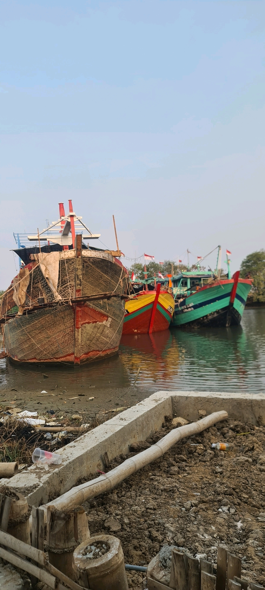 A scene in Juwana, Indonesia depicts several fishing boats moored in a body of water. The boats, varying in color and size, are lined up, some appearing weathered and covered with netting. The foreground shows a muddy bank littered with debris, including plastic bottles and construction materials. In the background, trees and a cloudy sky are visible. The water reflects the sky and the boats.