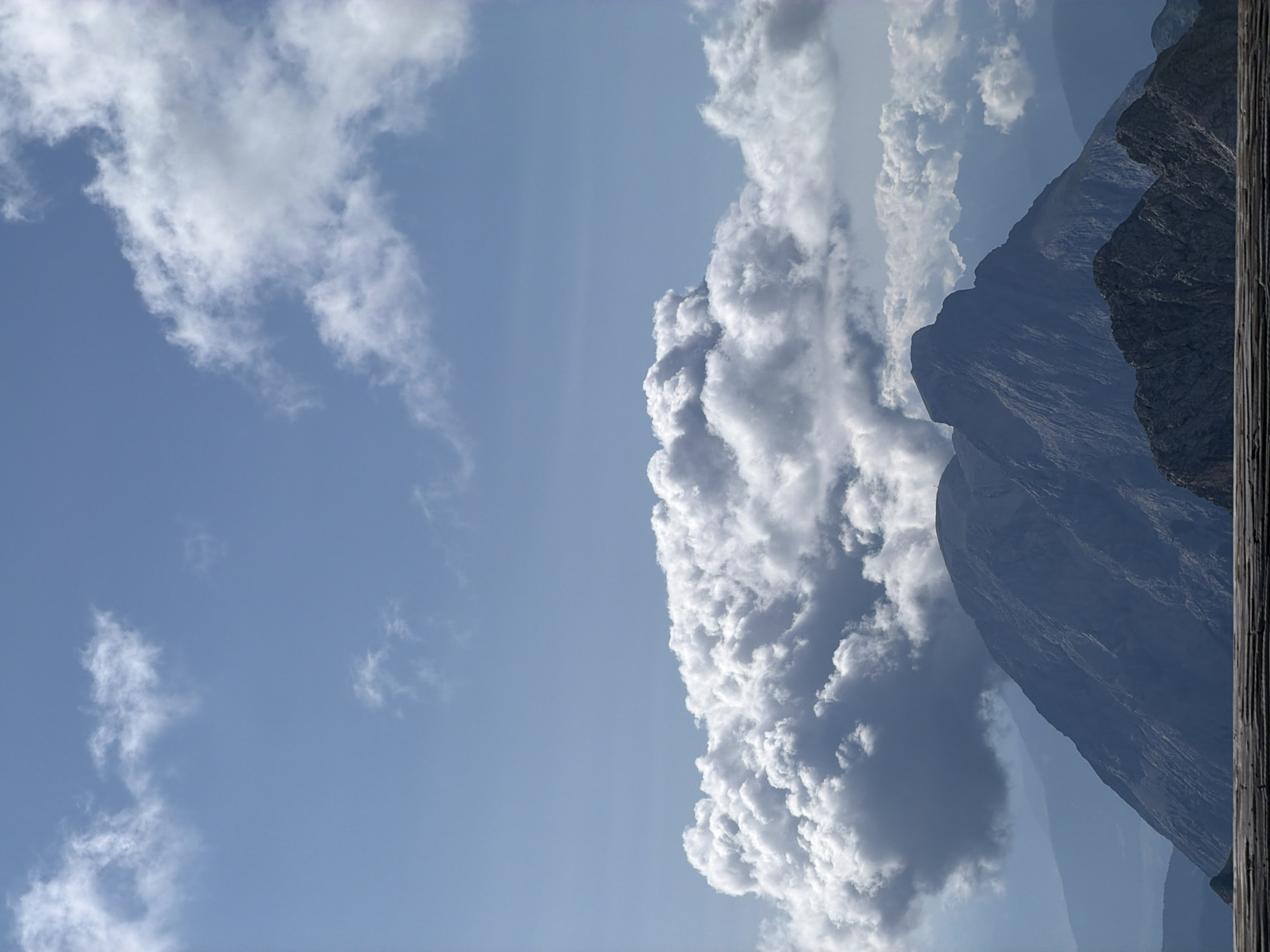 The image depicts a mountainous landscape under a partly cloudy sky. The foreground features a wooden railing, likely part of a viewing platform. Beyond the railing, a large, dark mountain dominates the mid-ground, with its rugged slopes casting shadows. Behind this primary peak, a series of successively lighter, mist-shrouded mountain ranges recede into the distance. Large, billowy white clouds are clustered in the sky, with some darker, more ominous clouds to the left, partially obscuring the upper reaches of the main mountain. The sky itself is a bright, clear blue.
While the image shows a mountain landscape, there is no specific indication or visual evidence within the image that places it in Garmisch-Partenkirchen, Germany. The scene is generic to mountainous regions. There are no people, actions, or interactions visible. The focus is solely on the natural environment.