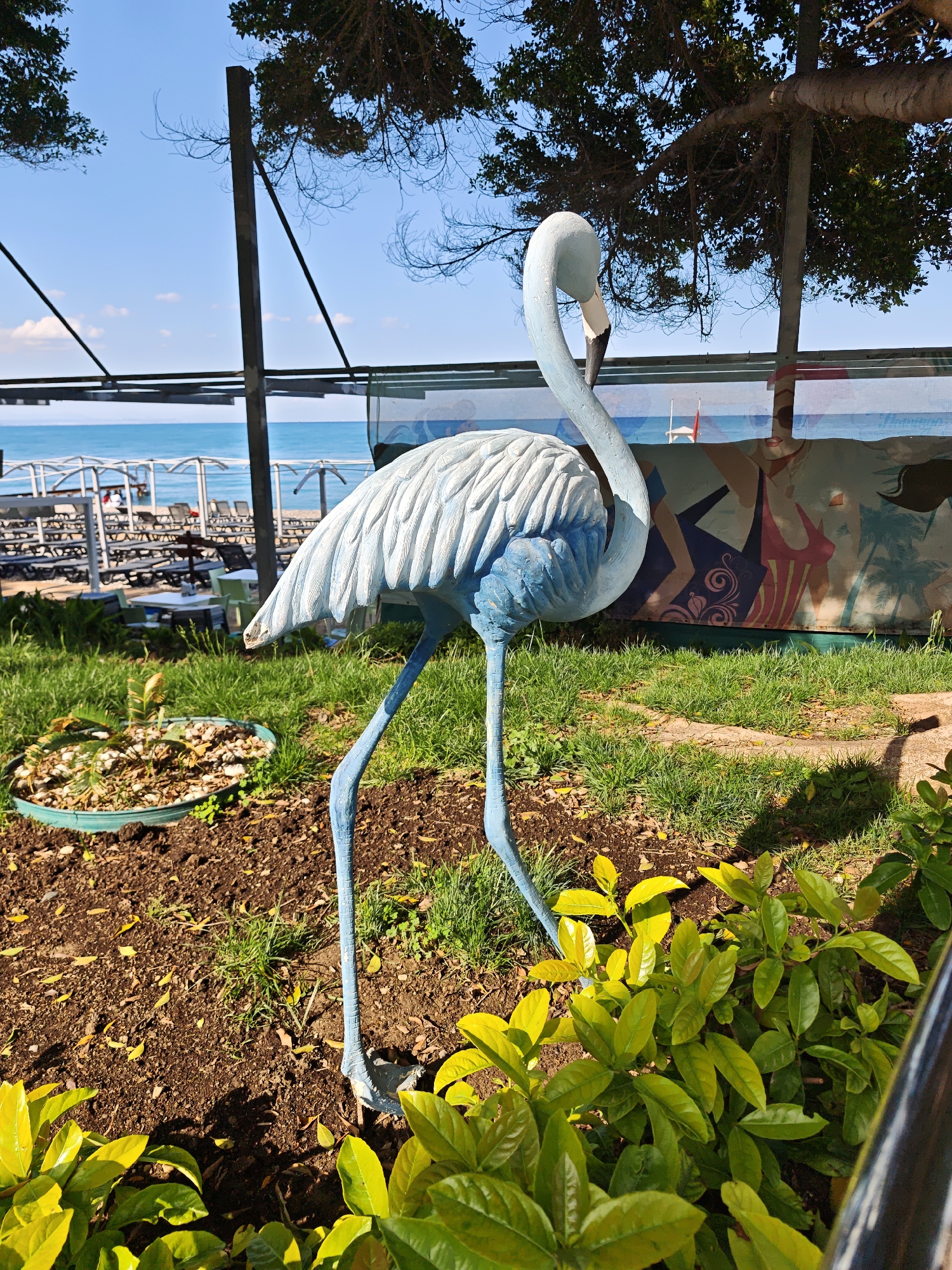 A blue and white flamingo statue stands on a patch of grass and dirt, with green plants around its base. Behind the statue, there's a view of a beach area with sun loungers and parasols. Further back, the blue sea is visible under a clear blue sky with a few white clouds. The scene is partially shaded by trees, and there's some infrastructure like metal poles and a structure with a mural visible in the background. This photo was taken in the city of Kemer, Turkey.