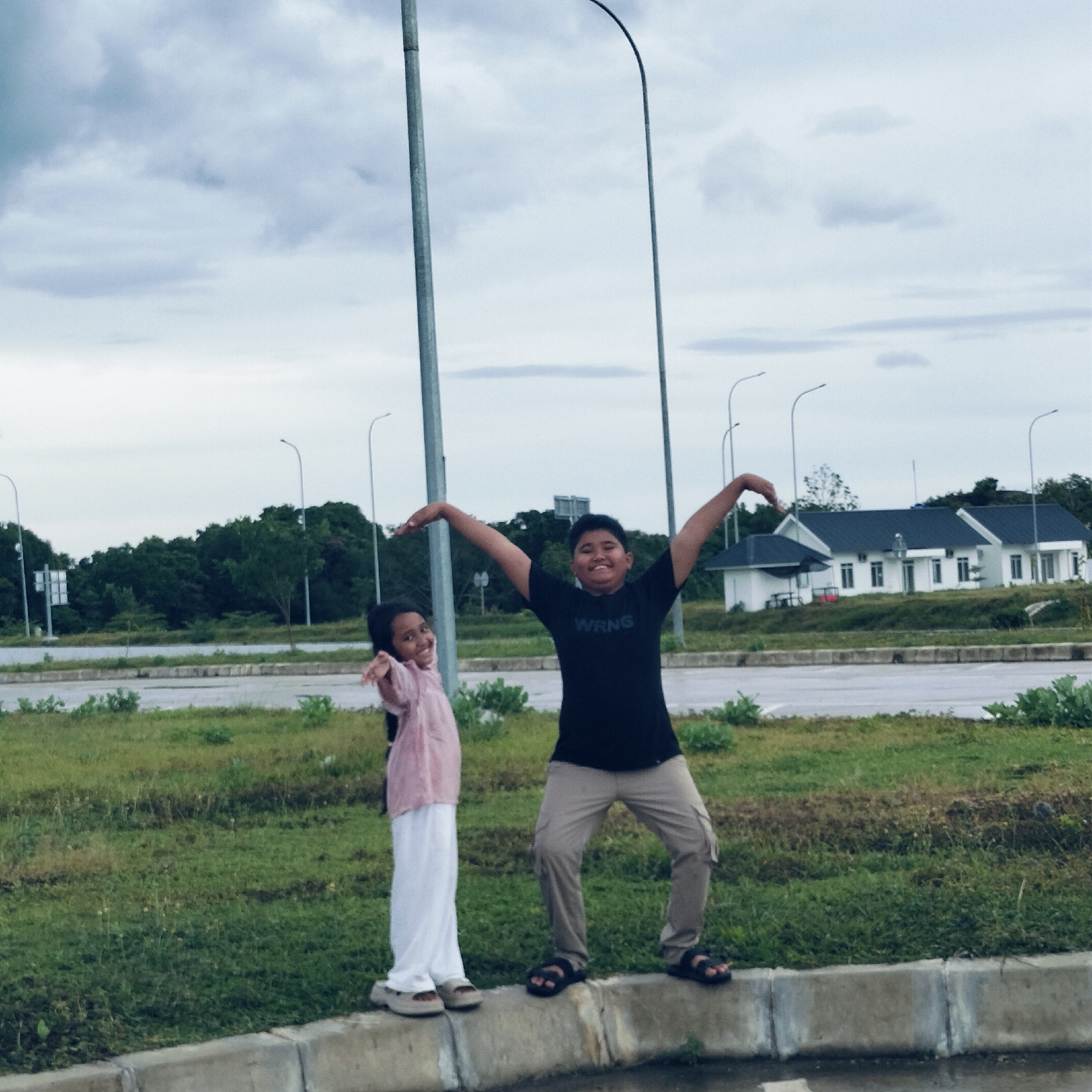 This outdoor photograph captures two children, a boy and a girl, standing on a concrete curb in what appears to be a developing or semi-urban area in Keureuweungkrueng, Indonesia.
The boy, on the right, wears a black t-shirt with the text "WRNG" prominently displayed, light brown cargo pants, and black sandals. He is striking a playful pose with both arms raised and outstretched, a wide, joyful smile on his face. The girl, positioned to his left, is dressed in a light pink long-sleeved top and white pants, wearing light-colored sandals. She looks towards the camera with a sweet smile, her left arm slightly bent and raised, mimicking a pose.
The children are positioned in front of a wide expanse of green grass, behind which lies a road. In the mid-ground, a dense line of trees stretches across the horizon. Several tall, slender streetlights with curved tops are visible, extending into the sky. Further in the background, to the right, there are a few white buildings with dark roofs.
The sky is overcast with soft, grey clouds, indicating a cloudy daytime setting. The overall atmosphere of the image is cheerful and lighthearted, primarily conveyed by the children's happy expressions and dynamic poses.