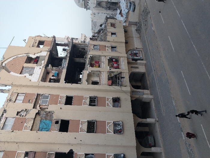 This image depicts a scene of extensive destruction in an urban environment, likely in Khan Yunis based on the provided context. The primary subject is a multi-story residential or commercial building that has suffered severe structural damage, with large sections of its facade and interior blown out or collapsed.
The building, which appears to be light beige, shows multiple floors with exposed rooms, missing walls, and visible debris. A significant portion of the building, especially towards the upper middle, appears charred and blackened, indicating fire damage. Despite the devastation, some signs of former habitation are visible, such as clothes hanging on railings or inside damaged rooms (red, blue, pink items), and a light blue tarp or fabric draped in one of the window openings. The building's architecture features decorative elements around some windows and arched entrances on the ground floor. A vehicle is partially visible parked under one of these archways.
In the foreground and to the right, a paved road stretches diagonally across the frame, featuring white lane markings. Three individuals are visible on the road: two people, dressed in dark clothing, appear to be walking or running away from the building, while a third person, wearing red, is crouching closer to the damaged structure.
In the background, beyond the damaged building, more urban structures are visible, including what appears to be a large, white domed building or mosque, suggesting a significant landmark. Other damaged buildings or piles of rubble can be seen further in the distance. The sky is clear and bright.
A small billboard or sign attached to the damaged building on the right side displays an image, possibly of a red tractor or agricultural equipment, with some visible Arabic text which appears to include "المزرعة" (Al-Mazra'a), meaning "The Farm." The overall impression is one of widespread devastation following conflict or a major incident.