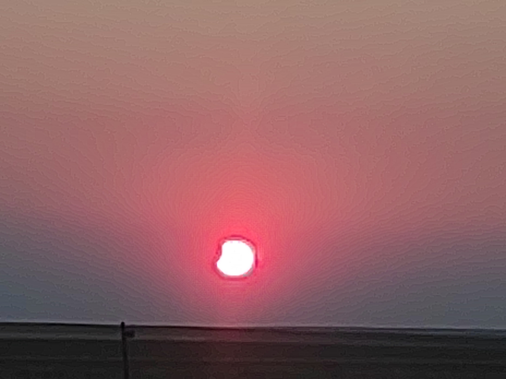 The image depicts a sunset over a flat, open landscape. The sky is filled with shades of pink and purple, with a bright, circular sun near the center, appearing red and partially obscured by atmospheric haze. The horizon line is low, with a dark, indistinct strip of land or water below the sky. A thin, vertical black object, possibly a pole or a distant silhouette of a person, is barely visible against the dark landscape near the bottom left edge of the frame. The overall impression is one of a serene, twilight scene in a rural or coastal environment. While the image is stated to be from Autillo de Campos, Spain, this information is not visually discernible from the photograph itself.