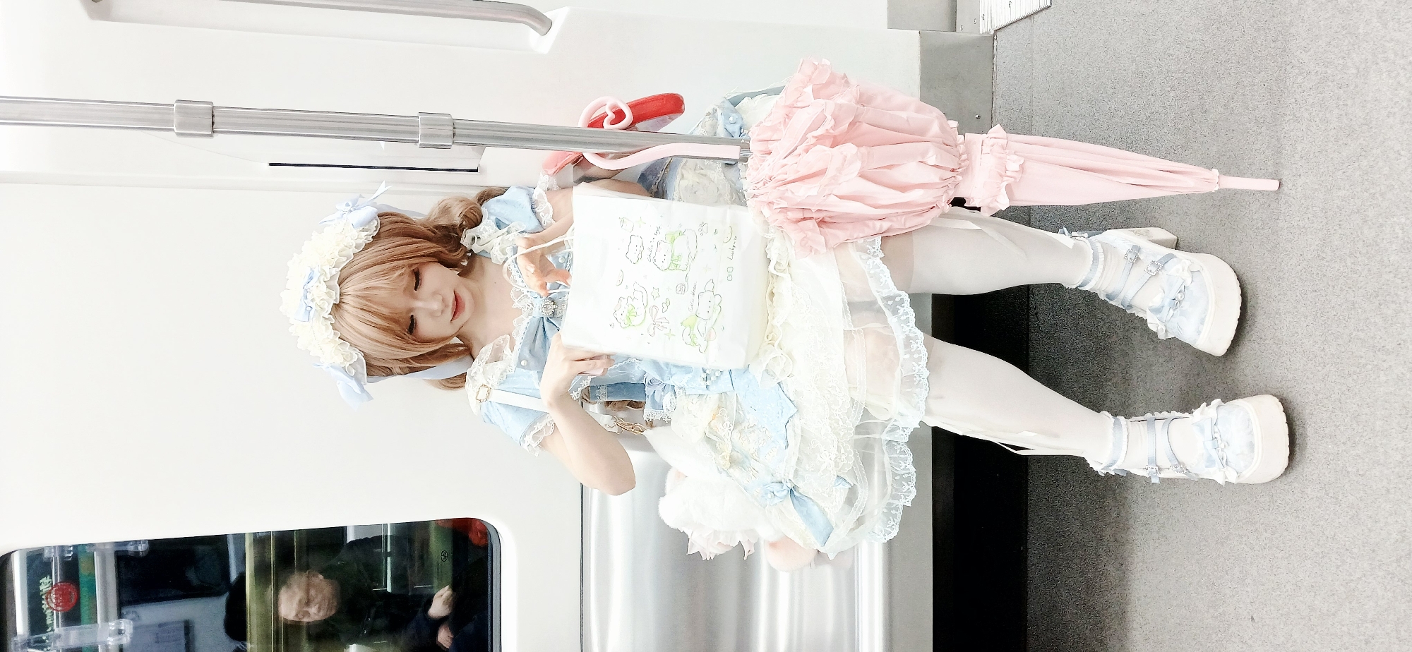 A young woman wearing a Lolita dress stands inside a subway car. She is wearing a light blue and white dress with lace detailing and a matching headband. She also has white tights and platform shoes. She is holding a white bag with cartoon characters on it. Next to her is a pink umbrella with ruffles. The background is a clean, modern subway car interior with metal handrails and a window showing people outside. The location is likely the city of Chongqing High-tech Zone, China, based on the style of the subway car.