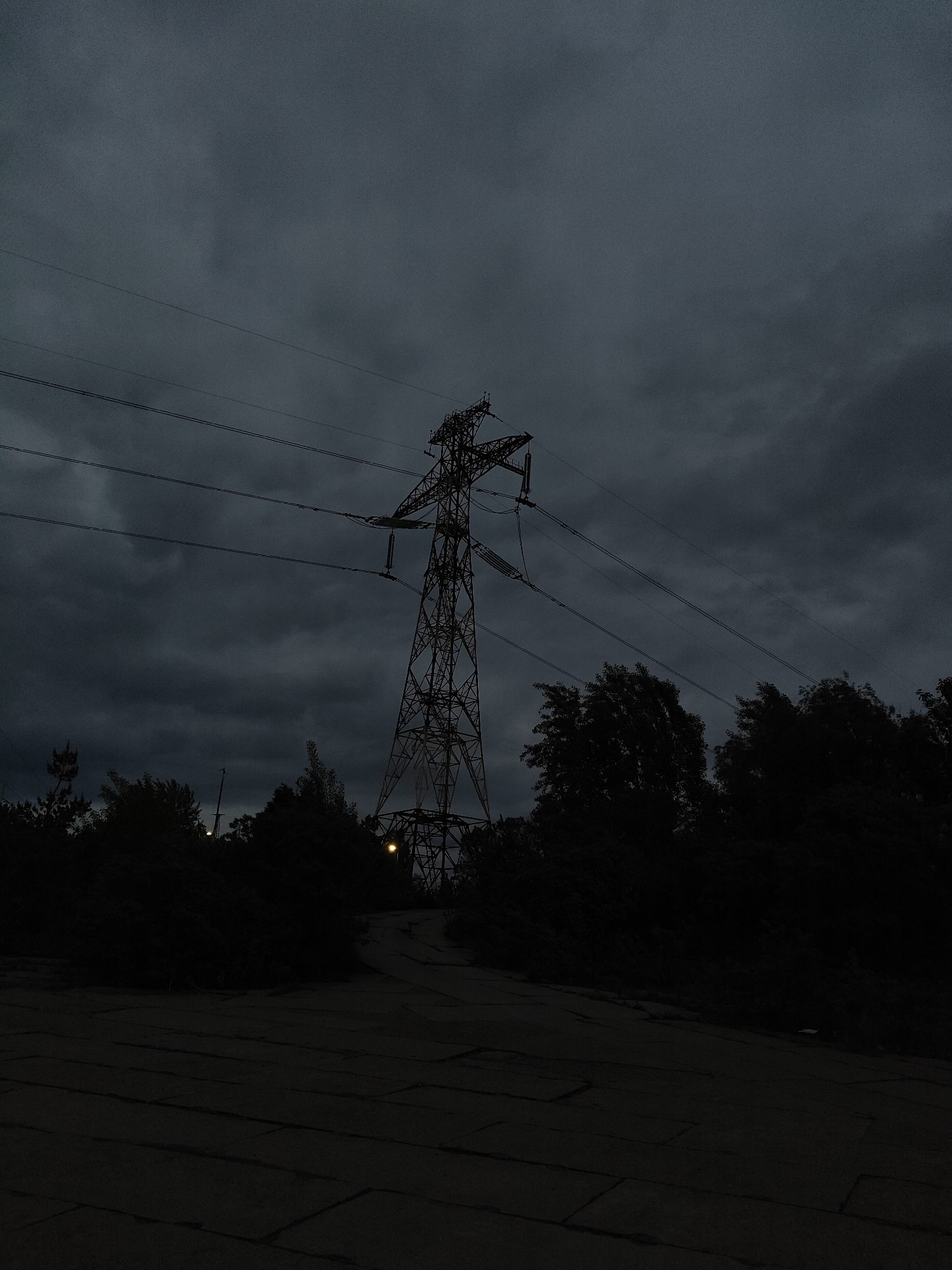 The image depicts an electrical transmission tower in Chaikovsky, Russia under a dark, overcast sky. The tower is silhouetted against the gloomy sky. Power lines extend from the tower. The surrounding area appears to be a combination of vegetation and a cracked concrete surface or roadway in the foreground. Two yellow lights are visible on the tower. The scene is dimly lit, suggesting either night or very low daylight conditions.