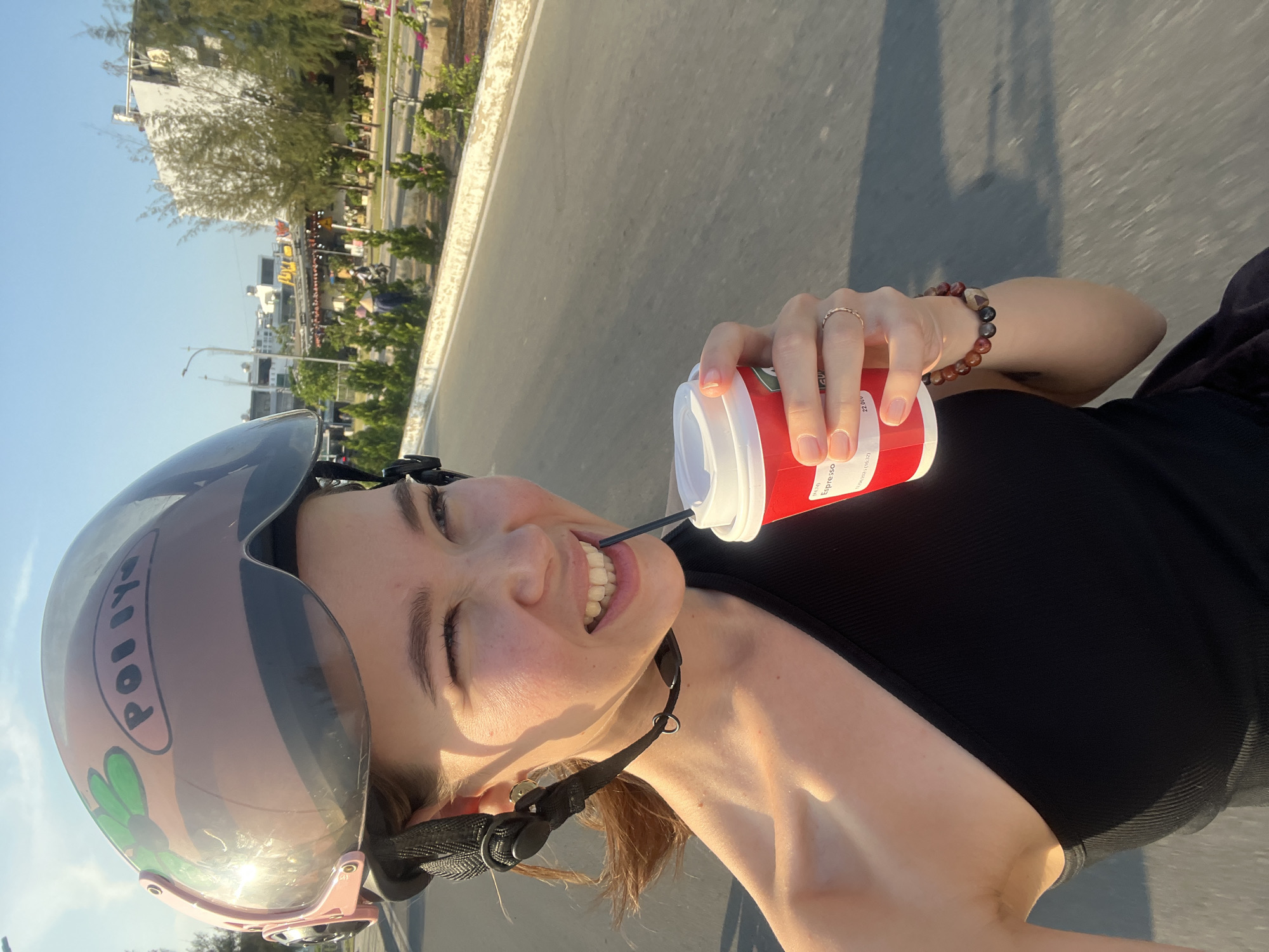 A young woman is captured in a dynamic selfie, smiling broadly with eyes crinkled in enjoyment, likely while riding a scooter or motorcycle in the Nam Natural Heritage Area Trang, Vietnam. She is looking directly at the camera.
She wears a light pink motorcycle helmet with a clear visor, which reflects strong sunlight. The helmet is personalized with the name "POLLY" and a green flower design. She is dressed in a black, ribbed tank top. Her right hand, adorned with a delicate gold ring and a beaded bracelet featuring brown and black stones, holds a red and white disposable cup. A black straw extends from the cup into her mouth, indicating she is drinking. The cup has "ESPRESSO" printed on its side, along with "22.0ev" and smaller text including "1/10 (3/10)" and a sequence starting "16:32".
The background shows a paved road or path under a clear, bright blue sky. Distant trees and buildings line the roadside, suggesting an urban or semi-urban environment. The strong sunlight and clear sky indicate a sunny day, likely during late afternoon or early morning, judging by the lighting and shadows. The overall image conveys a cheerful and active moment.