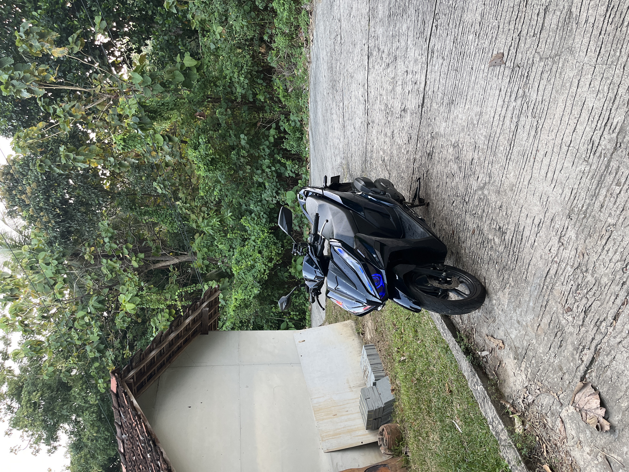 In the city of Legonipah, Indonesia, a black scooter with blue accents on its headlights is parked on a rough concrete surface. To the left of the scooter is a small, light-colored building with a terracotta tile roof. Next to the building, on the grassy ground, are several stacks of grey tiles and a terracotta pot. The background is filled with dense green trees and foliage, suggesting a natural, possibly rural or semi-rural setting. The sky is overcast.