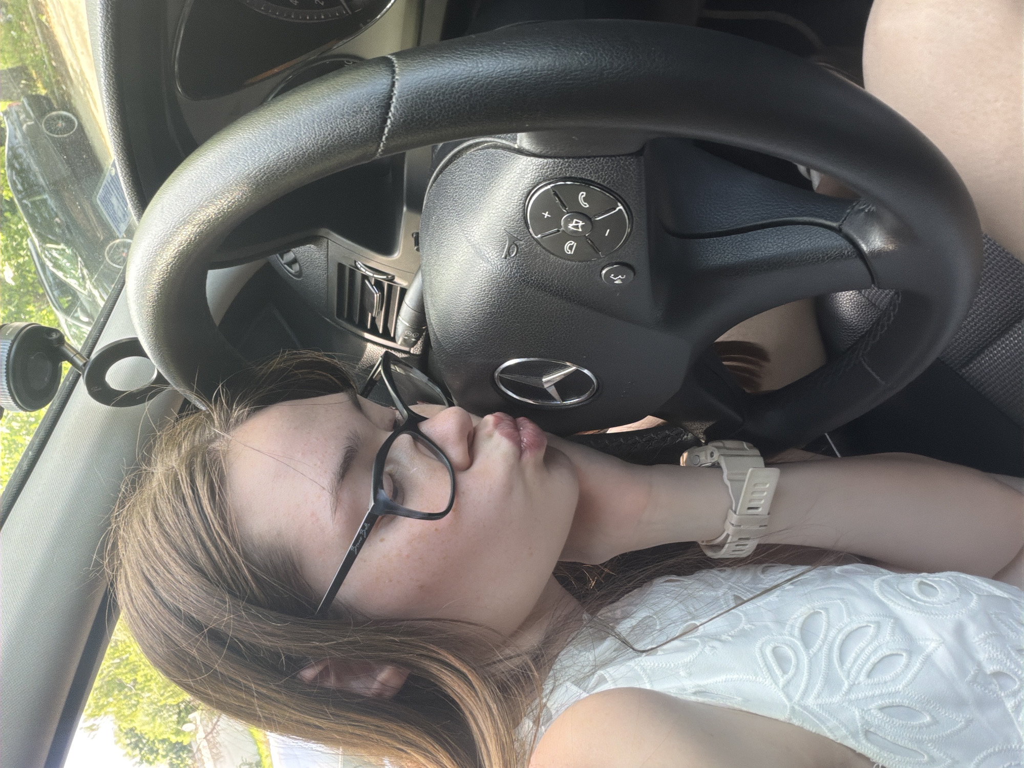 A young female is seated within the interior of a Mercedes-Benz vehicle, located in Varna, Bulgaria. She is captured in profile, leaning her left cheek on her left hand, while her lips are puckered in a kissing gesture towards the Mercedes-Benz logo centered on the steering wheel's airbag cover. The individual has long, light brown hair, wears dark-framed eyeglasses with "Brian Belle" visible on the left temple, and exhibits freckles on her nose and cheeks. She is dressed in a white, sleeveless top or dress featuring an intricate eyelet or embroidered pattern. A light-colored digital wristwatch is fastened to her left wrist.
The car's interior is dark, featuring a black, textured steering wheel with integrated circular multimedia and phone controls on the right spoke, beneath a small horn icon. Dashboard air vents are visible behind the steering wheel. Through the windshield, a blurred outdoor environment is visible, including green foliage from trees or bushes and a dark-colored sedan with prominent light-colored wheel rims. Sunlight illuminates portions of the car's interior and the individual. A light-colored overhead console is visible, along with a mounted device on the dashboard or windshield. The person's bare leg is visible below the steering wheel, indicating a short garment.