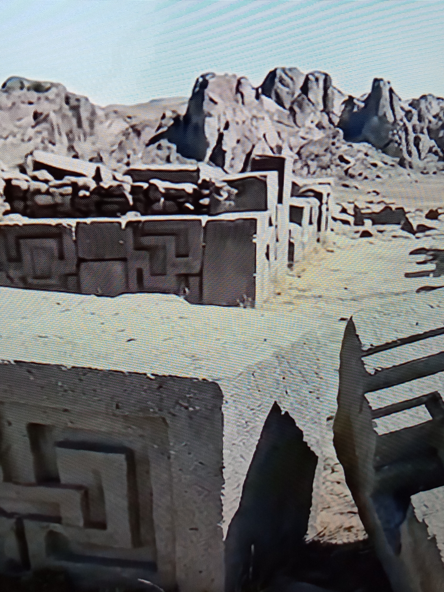The image shows ancient stone structures with intricate carvings in a rocky, arid landscape. The carvings appear to be geometric patterns. In the foreground, large, weathered stone blocks are visible, some with partially intact designs. In the background, a range of rugged, eroded mountains dominates the horizon under a clear, bright sky. The overall scene suggests an archaeological site or ancient ruins. The image does not contain any people or actions. There is no information to suggest the location is Brno, Czechia.