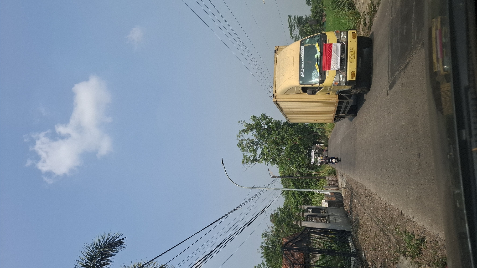 The image depicts a street scene in Sukasari, Indonesia. A yellow truck with a cargo container is traveling down a paved road, with the Indonesian flag displayed on its front. To the left of the road, there is a fence and a building with a tiled roof, bordered by vegetation. A street light is visible on the left side of the road. Further down the road, a person on a motorcycle is riding in the same direction as the truck. The sky above is clear blue with a few scattered white clouds. Green trees and vegetation line both sides of the road, with a rice paddy field visible on the right side. The scene appears to be captured during daylight.