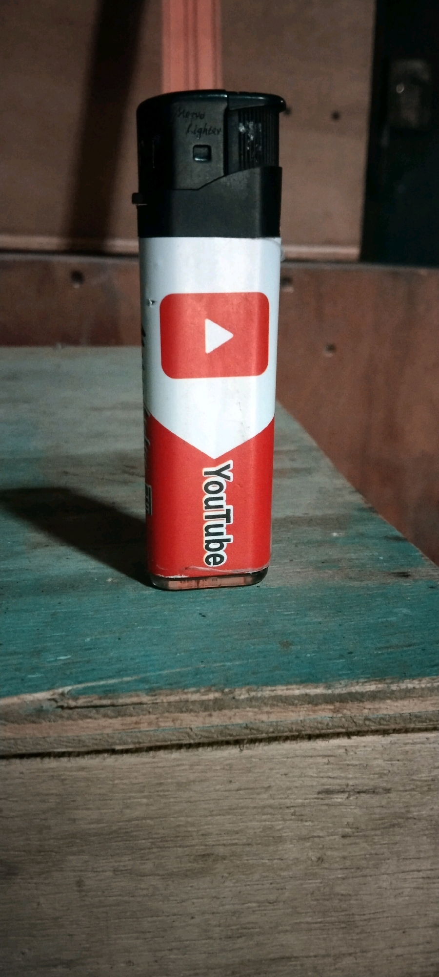 A close-up, eye-level, medium shot shows a single, black and white plastic cigarette lighter with a YouTube logo printed on its side. The lighter is standing upright on a turquoise-painted wooden surface. The background consists of out-of-focus wooden panels and a hint of a reddish vertical object. The lighting is dim, creating shadows on the surface and behind the lighter. The image is devoid of human presence. The scene does not provide any information to determine its location within the city of Wadang, Indonesia.