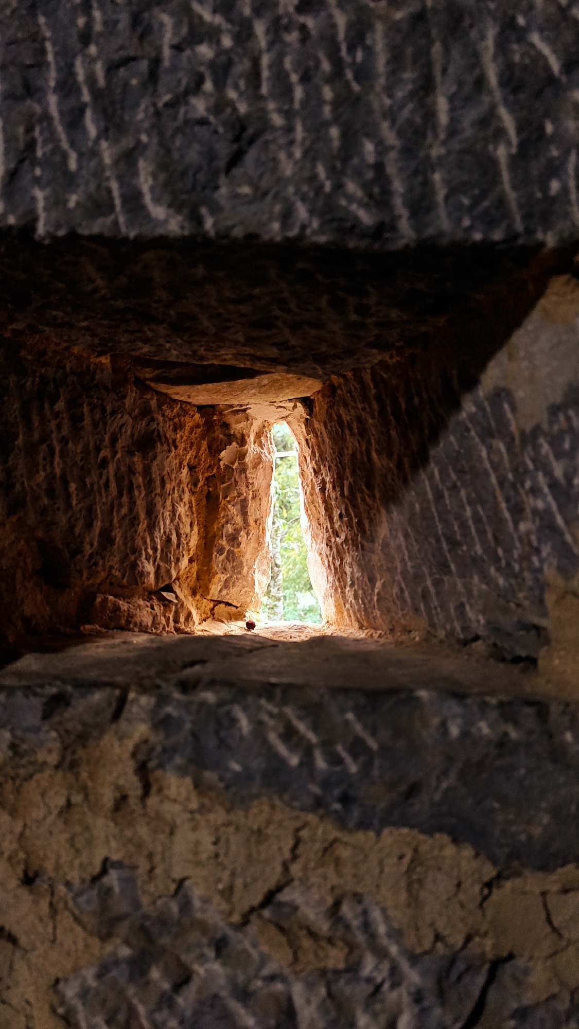 A narrow rectangular opening pierces a rough-hewn stone wall, revealing a glimpse of vegetation bathed in sunlight. The opening's interior is rough and uneven, appearing naturally formed, with an orange hue contrasting sharply with the dark gray stone of the wall. This vantage point is located in Sà Phìn Commune, Vietnam.
