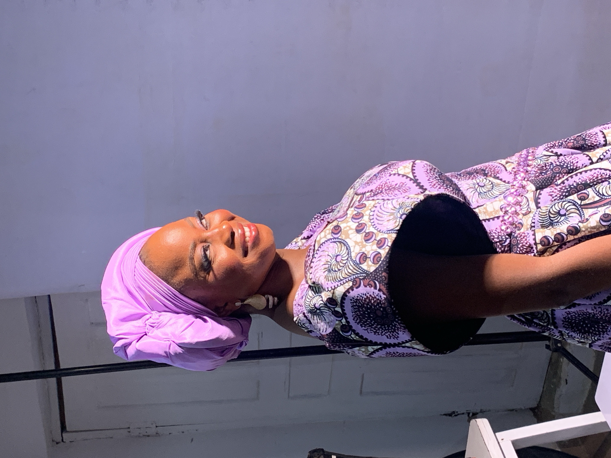 A woman with brown skin and a purple headwrap is turned slightly to the left, smiling and looking upwards. She is wearing a patterned dress in shades of purple, brown, and white. Her left shoulder is visible, and she has light-colored earrings. The background appears to be a room with a white or light gray wall and a dark horizontal bar. There is no information in the image to suggest the location is Jos, Nigeria.