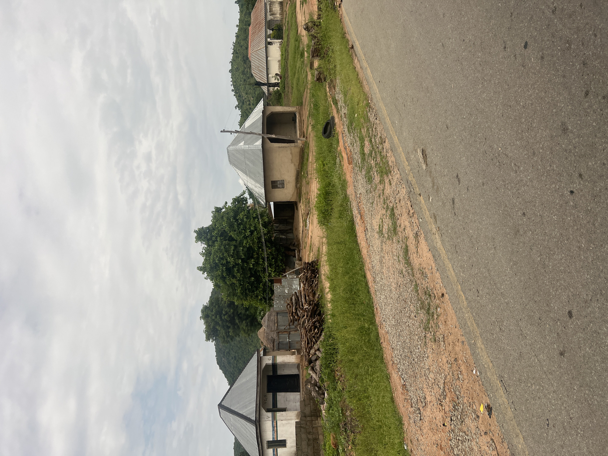 A moderately cloudy, overcast sky looms above a rural scene in Minda, Nigeria. In the foreground, a paved road runs diagonally across the frame from bottom right to top left, with a dirt and gravel shoulder. Beyond the shoulder, a patch of lush green grass leads to a collection of small buildings.
To the left, a white rectangular building with a dark, pyramid-shaped metal roof stands beside a smaller structure with a thatched roof. In front of these buildings, a substantial pile of cut logs lies on the dry earth. Further to the right, a small, mud-colored hut with a similar metal roof, open on one side, is situated in a sandy yard. A discarded black tire rests nearby.
Further back, another building with a red-tiled, gabled roof is visible. The landscape behind the buildings is characterized by rolling green hills covered in trees. The overall impression is one of a modest rural settlement.