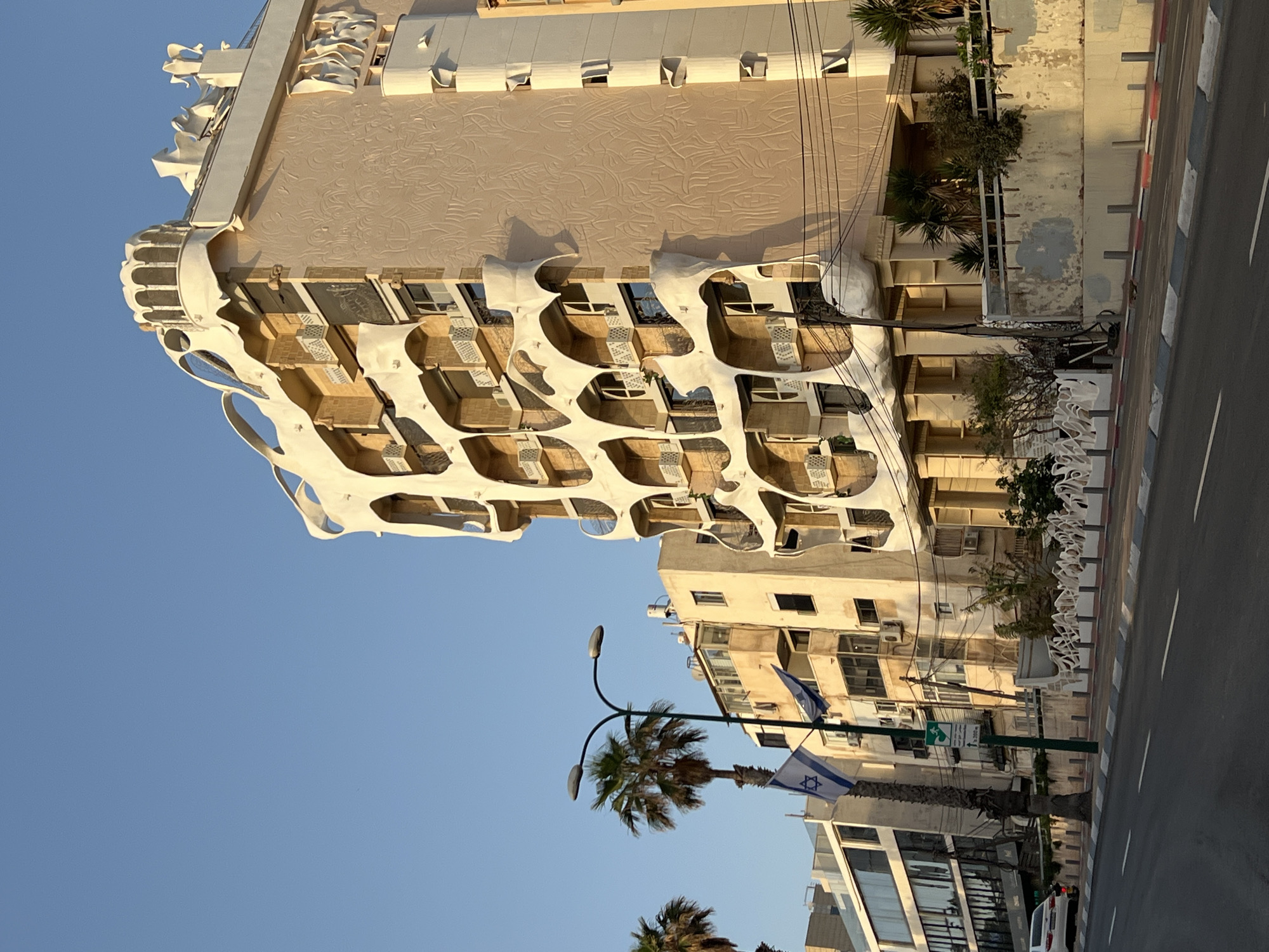 The image depicts a street scene in Tel Aviv, Israel, featuring an architecturally unique building with organic, bone-like forms surrounding its windows and balconies. The building has a light beige facade with a textured pattern on the upper portion. The street is lined with other buildings, palm trees, and streetlights. The Israeli flag is visible on a pole near a traffic sign. A road with white lane markings and a striped curb is on the right side of the image. The sky is clear and blue.