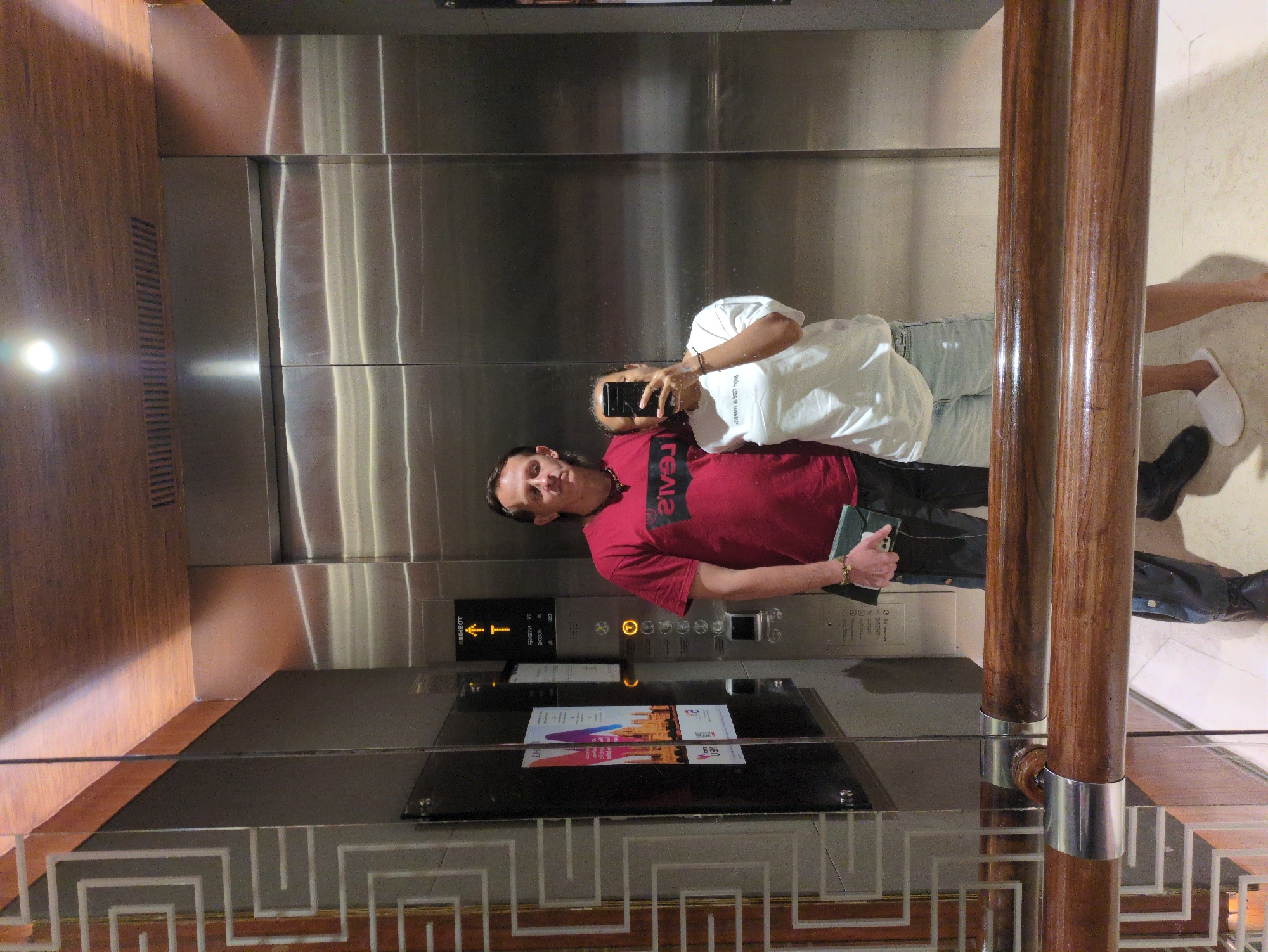 A man and a woman stand in an elevator, taking a mirror selfie. The man is wearing a red t-shirt and dark pants, and he is holding a green folder. The woman is wearing a white t-shirt and denim shorts, and she is holding a smartphone to take the picture. The elevator has stainless steel walls and a wooden ceiling. The floor appears to be tiled. The elevator doors are closed. The location is not in the city of Agra, India.