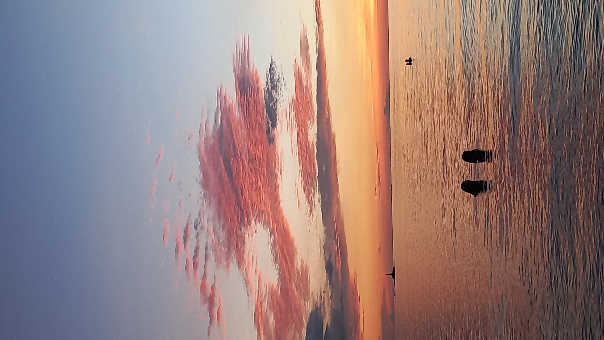 This image depicts a serene sunset scene over the ocean. In the foreground, two people are partially submerged in the water, with only their heads visible. Further in the distance, another person is seen in the water, and a small lighthouse stands on a rocky outcrop on the horizon. The sky is painted with hues of orange, pink, and blue, with scattered clouds reflecting the sunset colors. The water's surface is calm, with gentle ripples. The overall atmosphere is tranquil and picturesque. It is difficult to determine the exact location within Tambon Ko Phangan, Thailand based solely on this image.