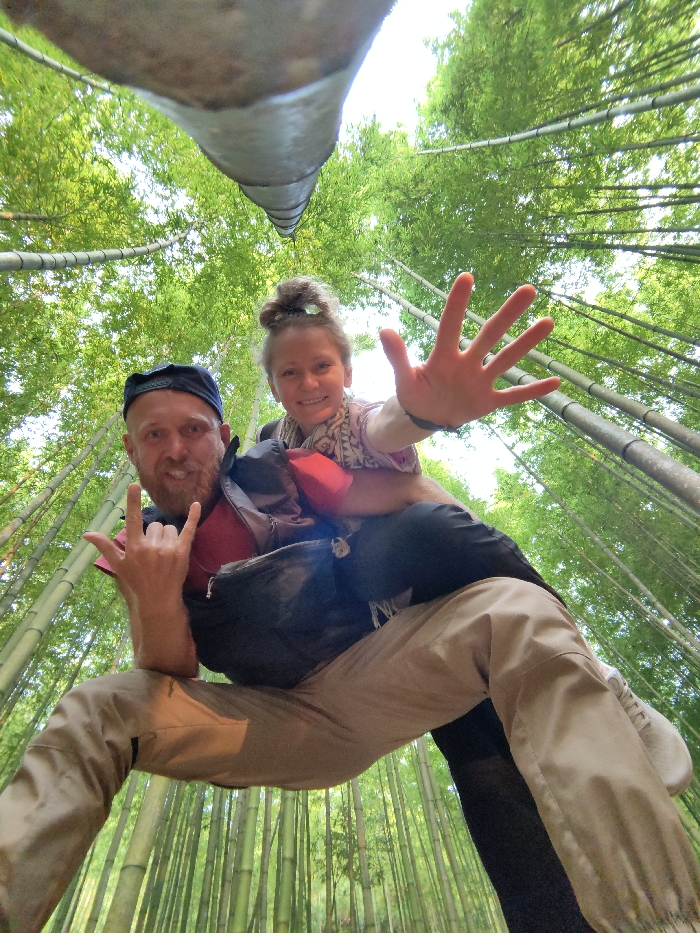 An adult male and a young female child are depicted in a low-angle photograph within a dense bamboo forest, identified as Mù Cang Chải, Vietnam. The light-skinned adult male, estimated to be in his 30s-40s, with a beard and a black baseball cap worn backward, squats in the foreground, facing the camera with a smile. He wears a dark shirt and beige cargo-style pants, with white sneakers visible. His left hand is raised, forming a "rock on" gesture. A light-skinned female child, appearing to be 5-10 years old, with her hair in a bun, is seated on the man's back, supported by a dark carrier or backpack with a red accent visible on the man's shoulder. The child also smiles at the camera, extending her right hand forward with fingers spread. The background consists entirely of numerous tall, slender green bamboo stalks extending upwards, with visible green foliage at the canopy level, obscuring most of the sky. A prominent, thick bamboo stalk traverses the upper left portion of the frame, close to the camera.