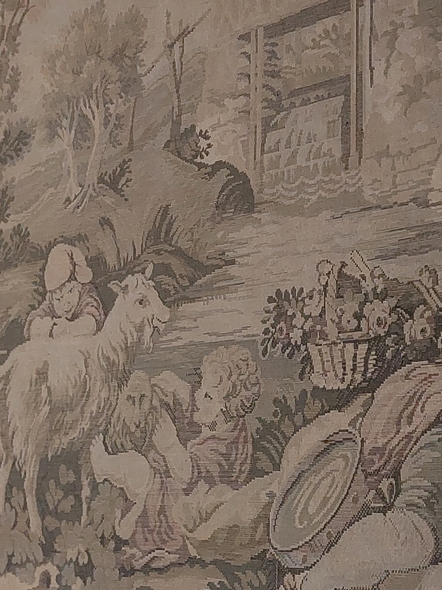 The image is a close-up of a tapestry depicting a pastoral scene. In the foreground, a child wearing a cap is seated on the back of a goat. The child is looking down, appearing to interact with something near the goat's head. To the right, another child, with curly hair, is seated and reaching out towards a basket filled with flowers. Behind the children and the goat, a textured wall, possibly part of a building or structure, is visible. The background features trees and a hilly landscape, rendered in muted tones. The tapestry displays a detailed, somewhat stylized representation of rural life. This tapestry is located in the city of Cesa, Italy.