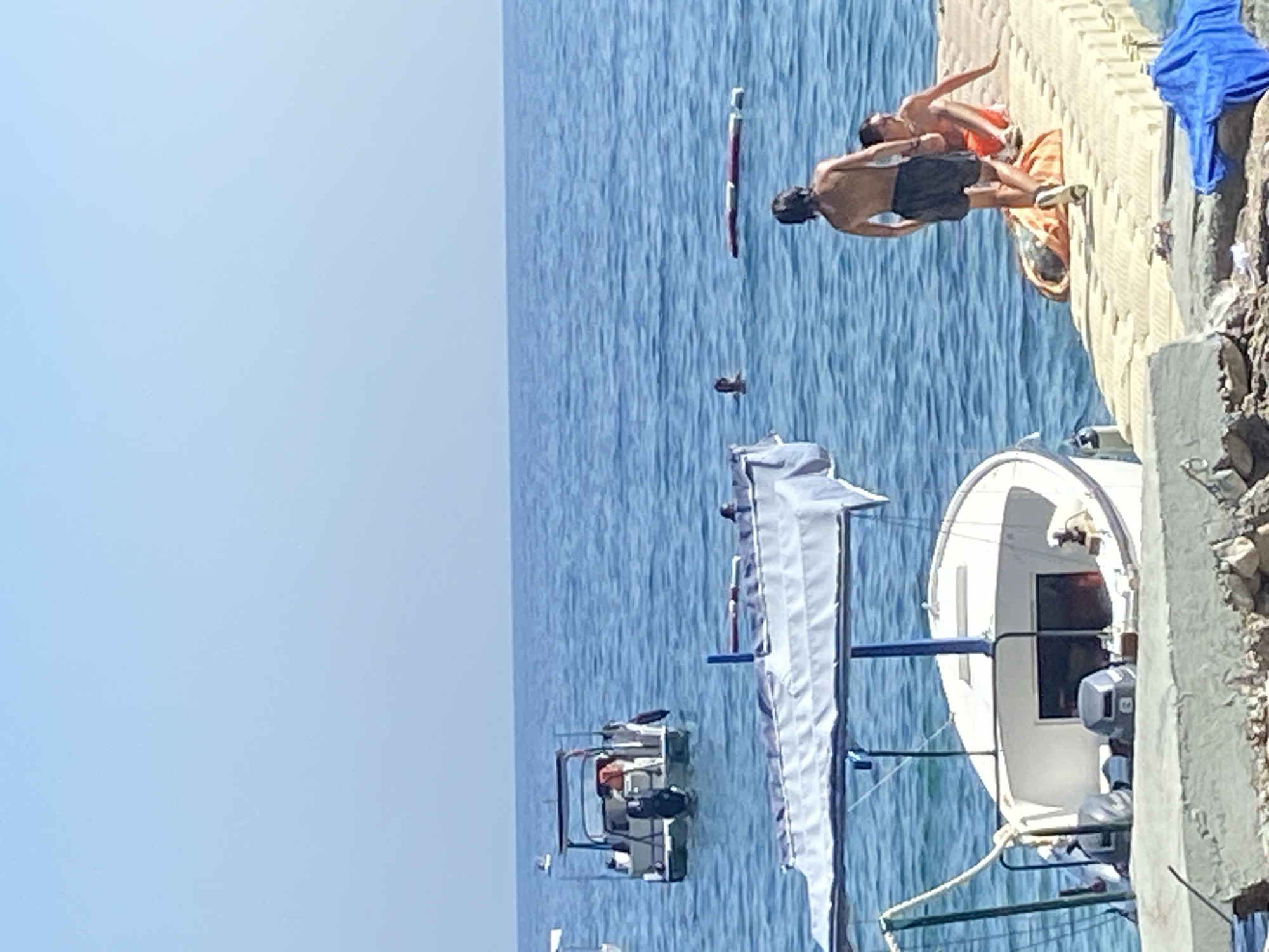 In Prijevor, Montenegro, on a clear, sunny day, a tranquil scene unfolds at the water's edge. The deep blue sea stretches out under a pale blue sky, dotted with a few boats. In the foreground, a white boat with a blue canopy is docked at a concrete pier, its two outboard motors visible. Further out, a small motorboat, also equipped with an outboard motor, floats on the calm water. Two individuals are on the pier. One, a young male wearing black shorts, is walking away from the camera. Another, also a young male, is seated on the pier wearing orange shorts. In the water, a person is visible swimming. The scene captures a serene moment of leisure by the sea.