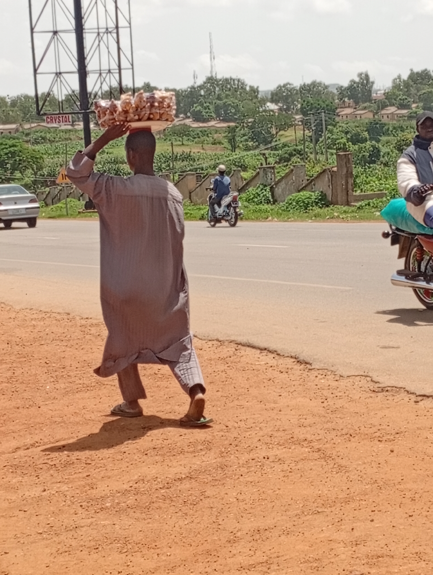 A man, wearing a long grey tunic and trousers, walks along a reddish-brown dirt road, carrying a large tray of food items wrapped in clear plastic on his head. The man is in motion, with his right leg forward and his left leg bent. His shadow stretches out behind him. In the background, a paved road runs parallel to the dirt path, with a few vehicles visible, including a silver car on the left and two motorcycles on the right. One motorcycle carries a passenger wearing a blue jacket, while the other is ridden by a person wearing a white and black jacket and a black cap, with a green bag secured behind them. Beyond the road, there is a green, overgrown landscape with scattered buildings and trees, and a large metal structure that appears to be part of a billboard with the word "CRYSTAL" visible. The sky is overcast. The scene is set in Rafin Par, Nigeria.