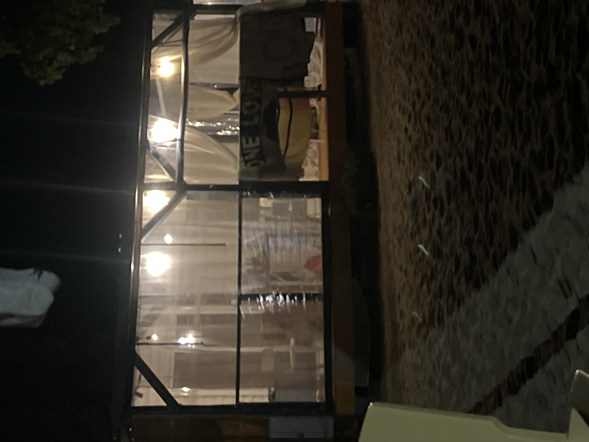 The image shows a beachside structure at night. The structure appears to be a covered seating area or bar with a wooden frame and transparent sheeting. Inside, there are lights and draped curtains. A banner with "ONE LOVE" written on it hangs from the front of the structure. There are also what appear to be towels draped over the railing. The ground in front of the structure is sandy, and small, glowing objects are scattered across it, possibly from a light display or glow sticks. A portion of a light-colored chair or table is visible in the lower left corner of the image. The lighting is dim, suggesting it is nighttime. The scene is consistent with an outdoor establishment on a beach in Sozopol, Bulgaria.