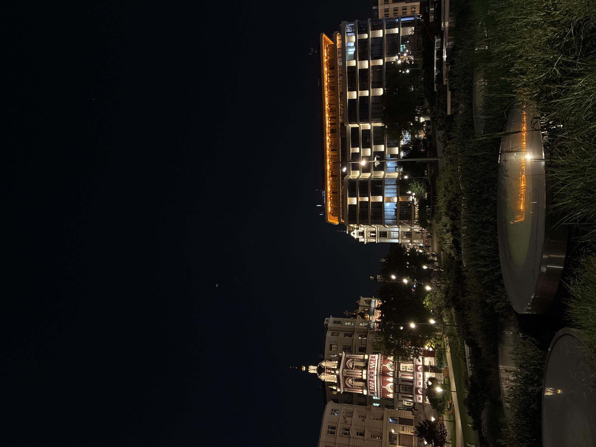 The image depicts a nighttime urban scene in Moscow, Russia. Dominating the view on the right side is a modern, multi-story building illuminated with orange and white lights. Adjacent to it is a park with greenery and reflective, circular installations. On the lower right, more traditional architecture is visible, with buildings featuring white and red facades and illuminated windows. The left side of the image is dark, with a clear night sky.