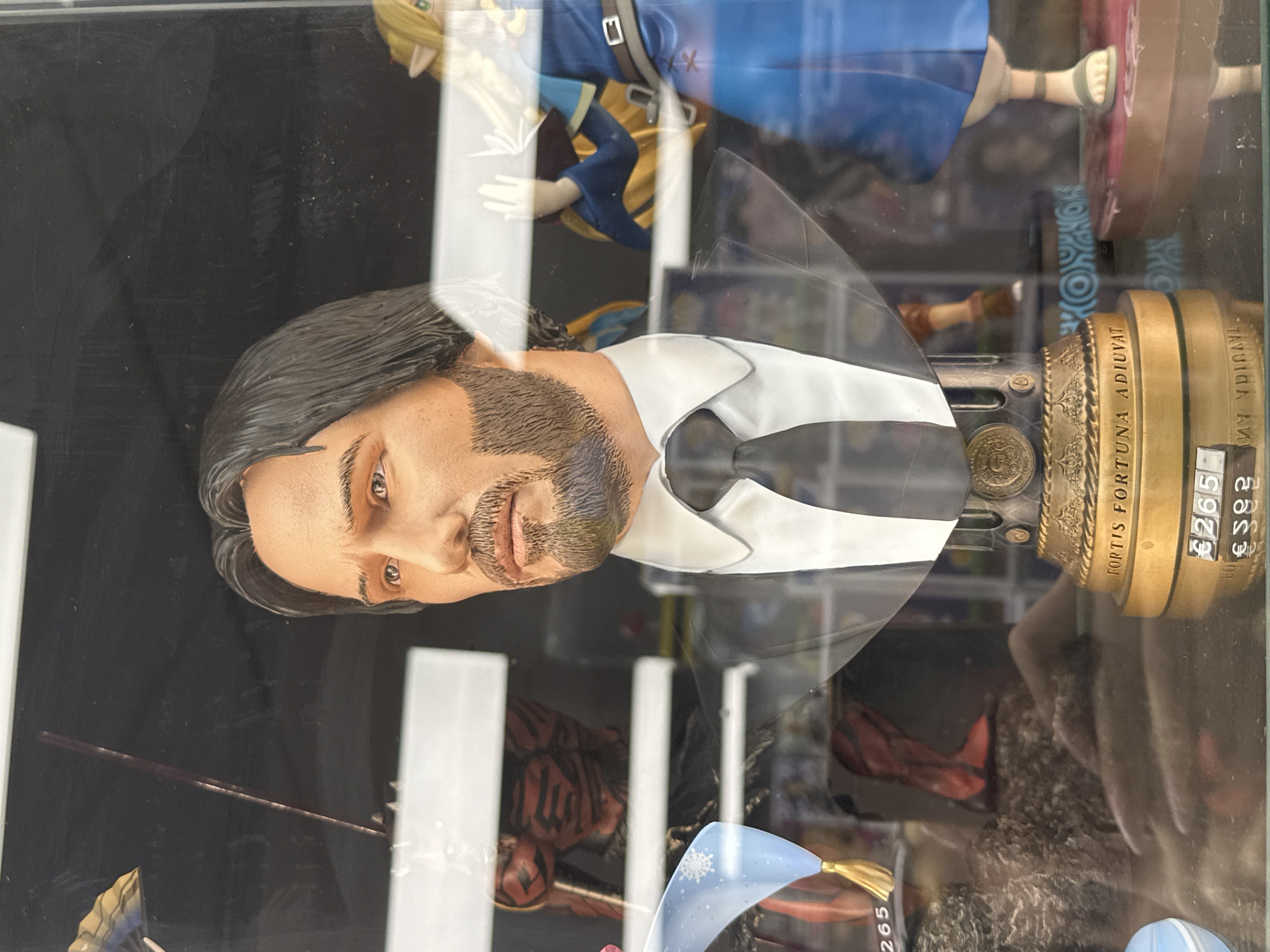 The image is a close-up, eye-level, vertical shot of a bust of a man, likely a collectible figurine, displayed in a shop window. The bust depicts a man with dark hair, a beard, and a stern expression, wearing a white shirt and a dark tie. The bust is mounted on a golden base with decorative elements and text that reads "FORTIS FORTUNA ADIU VAT".
Behind the bust, other figurines are visible, including a figure with red and black striped armor on the left and a female figure in a blue and gold outfit on the right, which resembles the character Link from The Legend of Zelda series. Reflections on the glass suggest the setting is indoors, possibly a toy or collectible shop. The price tag "€265" is visible on the base of the bust and another tag below it shows "€592". The overall lighting is somewhat dim, with the main subject well-lit.
Based on the presence of the price tag with the Euro symbol and the text "Luxembourg" visible on a label in the lower right corner of the image, the location is a shop in Luxembourg City, Luxembourg.
