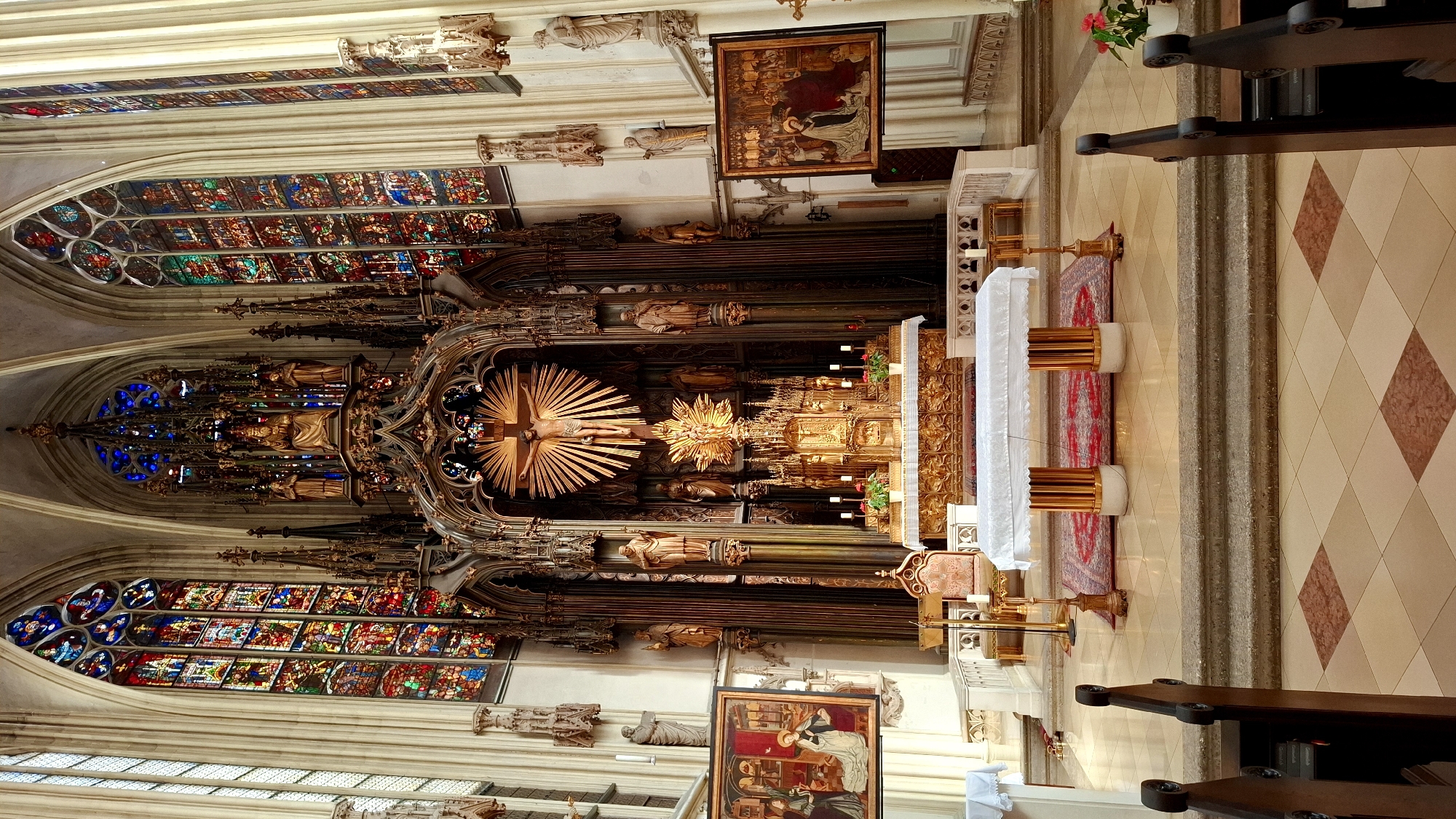 This is a detailed interior view of St. Stephen's Cathedral in Vienna, Austria, focusing on the high altar. The cathedral's Gothic architecture is evident with soaring ribbed vaults, pointed arches, and ornate decorative elements. Dominating the center is a magnificent, multi-tiered altar made of dark wood and gilded accents. At the pinnacle of the altar is a large crucifix depicting Jesus Christ, with rays of light radiating outwards. Below the crucifix, a golden tabernacle or reliquary is visible, adorned with intricate carvings and small candles in front of it.
Flanking the altar are tall, slender columns and niches containing statues of religious figures. To the left and right of the altar, large stained-glass windows with vibrant colors depicting biblical scenes are prominent. On the left wall, a framed painting hangs, depicting a religious scene with figures in historical attire. On the right wall, another similar framed painting is visible.
In front of the altar is a white-covered altar table, supported by golden cylindrical columns and resting on a patterned rug. The floor in the foreground is tiled in a diamond pattern with beige and reddish-brown tiles. Wooden pews are visible in the lower corners of the image, indicating the seating area for the congregation. The overall impression is one of solemnity, grandeur, and historical significance.