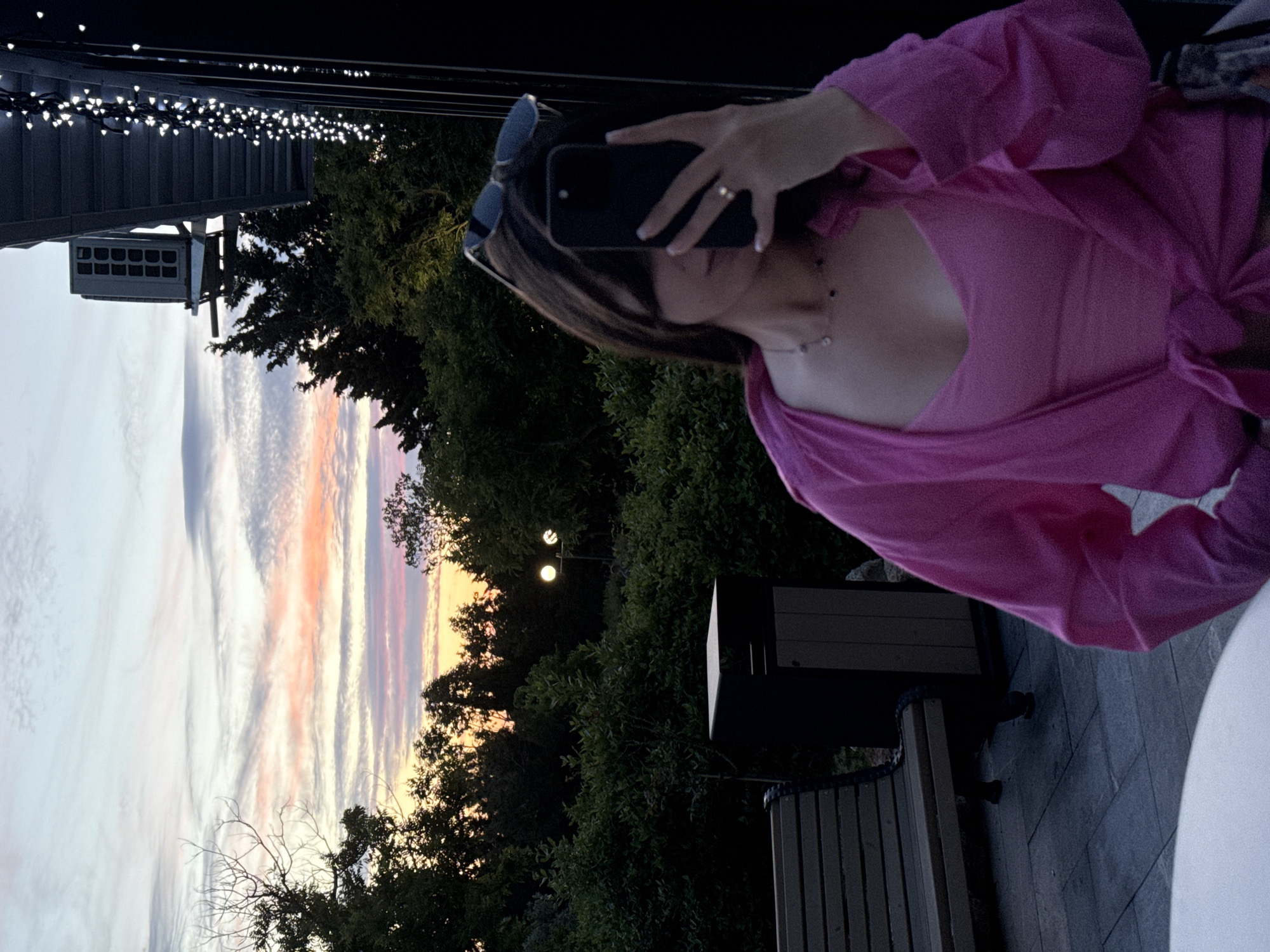 A young woman with dark hair takes a selfie in front of a sunset, her face partially obscured by her phone and hair, sunglasses perched on her head. She wears a pink tank top and a pink open-front shirt tied at the waist. A necklace with dark beads adorns her neck, and a ring is visible on her finger. The background reveals a tree-lined landscape with a bench, trash can, and outdoor lighting. In the upper right corner, an air conditioning unit is visible on a building. The sky displays a gradient of colors from pale blue to pink and orange, indicative of dusk.