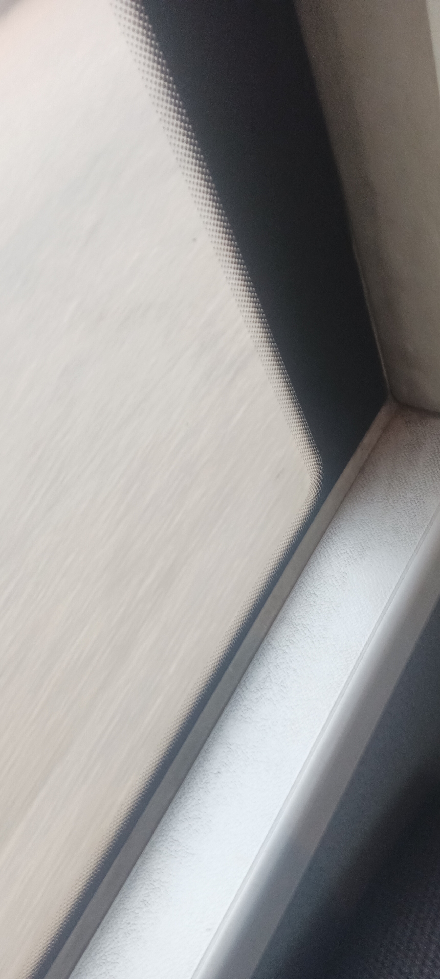 The image provides an abstract, close-up view from inside a moving vehicle, located in Gura Top, Nigeria. The primary subject is the interior of a vehicle, likely a car or bus, seen through a window. The left and central portions of the frame are dominated by a broad, light-colored expanse, which is heavily blurred due to motion, suggesting an external landscape or sky.
Along the upper right edge, a distinct pattern of small, dark dots is visible, characteristic of a frit band often found on vehicle windshields or windows. This dotted pattern acts as a transition zone between the blurred exterior and the vehicle's interior structure. To the right, part of the vehicle's interior is visible, featuring a darker, shadowed section in the top right corner. Below this, a lighter, textured surface, possibly part of the dashboard or window frame, is prominent, with its corner clearly defined.
The significant motion blur of the outside view indicates that the vehicle is actively traveling. The lighting suggests it is daytime. No people are visible in the frame, and there is no discernible text.