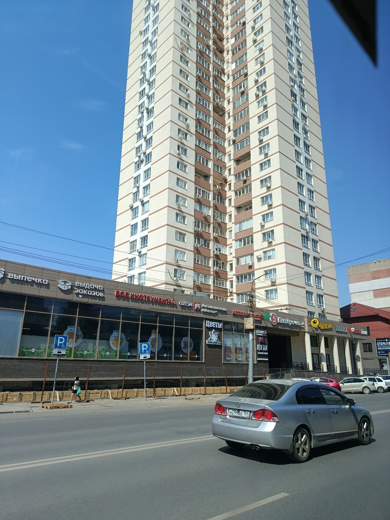 The image captures a street scene in Saratov, Russia, dominated by a tall, modern residential building. The building, with its beige and brown facade, features numerous windows and air conditioning units on each floor. Below the residential structure, a commercial strip is visible, with signs for businesses such as "Выпечка" (Baking), "Выдача заказов" (Order pickup), "ВСЕ ИНСТРУМЕНТЫ" (All tools), and "OZON". The storefronts have large glass windows, some displaying graphics and parking signs with the letter "P".
In the foreground, a silver sedan is driving on a multi-lane asphalt road. The car's license plate reads "H299BE 164". To the left of the road, a sidewalk is partially visible, with a woman walking and carrying shopping bags. Construction materials, including wooden planks and pallets, are present along the sidewalk. The sky above is clear and blue. Another building, partially obscured, can be seen to the right of the main residential tower.