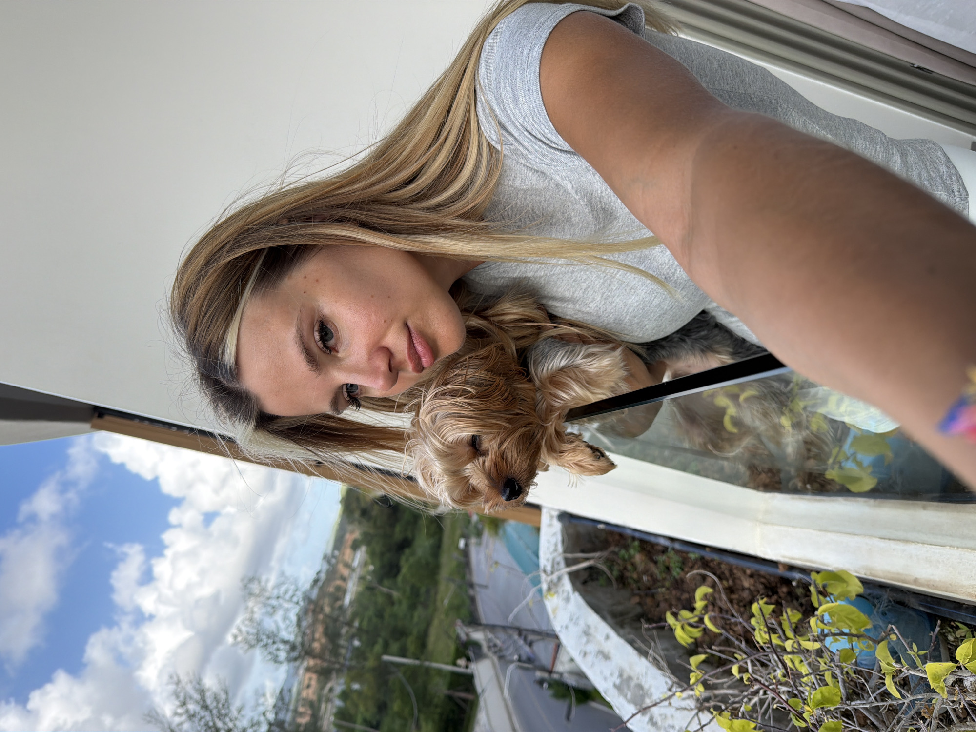 A woman with blonde hair and a grey t-shirt takes a selfie with her Yorkshire terrier on a balcony in Choeng Thale, Thailand. The dog is perched on the balcony railing, looking towards the camera with its eyes partially closed. The background shows a green, hilly landscape with a road winding through it, under a cloudy blue sky. The balcony has planters with foliage.