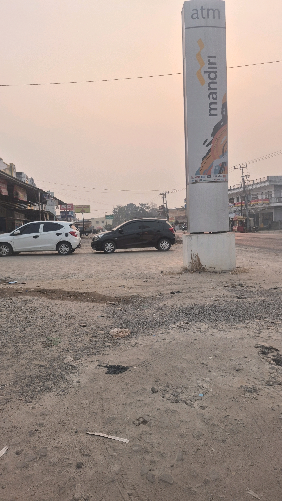 In Putak, Indonesia, a street scene unfolds under a hazy sky. A prominent ATM sign for "Mandiri" stands tall, its vertical format adorned with the bank's logo and artwork. Below the logo, various credit card logos are visible, including Visa and Mastercard. In the foreground, a dirt and gravel lot stretches out, marked with tire tracks from vehicles. Two cars are parked on the paved road: a white hatchback on the left and a black hatchback on the right, positioned at an angle as if in a parking lot. Behind them, the street continues, lined with buildings that appear to be shops or businesses. The overall atmosphere is that of a commercial area on a somewhat overcast or smoggy day.