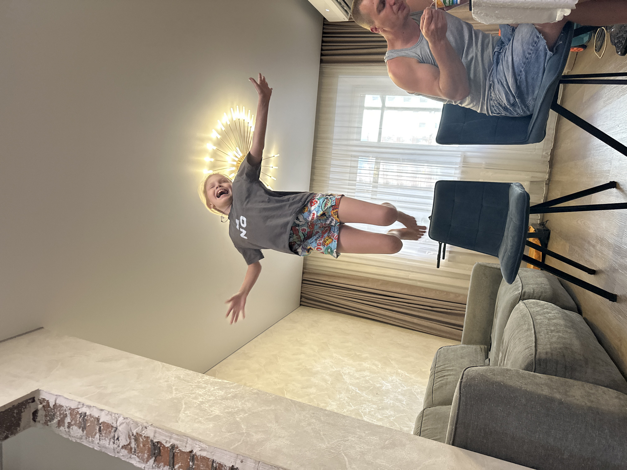 In this vertically oriented image taken indoors, a young girl with blonde hair is captured mid-air, appearing to jump or float. She is wearing a grey t-shirt with the letters "ON" visible in white, and brightly colored shorts with a comic book-like pattern. Her mouth is wide open in a joyful expression, and her arms are outstretched.
Behind her, a man is seated on a dark blue chair, wearing a grey tank top and denim shorts. He is looking downwards and appears to be holding something small in his hand, possibly a piece of food or a small object.
The scene is set in a room with light-colored walls. A large, ornate light fixture with many thin, bright bulbs is mounted on the ceiling, resembling a dandelion or starburst. There is a window with sheer curtains in the background, suggesting daylight filtering into the room. The floor is tiled with a light, marble-like pattern. To the right, a grey sofa is visible. The overall atmosphere suggests a casual, domestic setting, possibly a living room or dining area. The emotions conveyed are primarily joy and excitement from the girl, and a more subdued, observational demeanor from the man. No specific time of day or weather conditions are explicitly discernible beyond the presence of natural light.