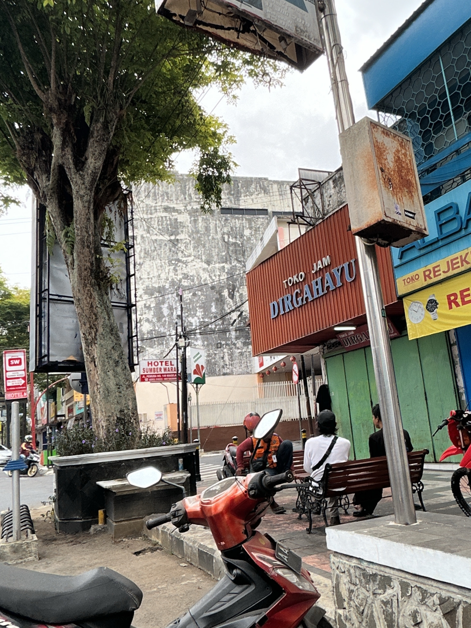 The image depicts a street scene in Magelang, Indonesia. In the foreground, a red and black motorcycle is parked. To the right, two men are seated on a dark brown bench, facing away from the camera. One man wears a white shirt with a black strap across his back and a black cap, while the other wears a black shirt. Behind them, a building with green shutters is visible. A sign above the shutters reads "TOKO JAM DIRGAHAYU" in orange and blue lettering. To the right of this sign, another sign with a yellow background displays an image of a watch and reads "TOKO REJEKI". Further to the right, a blue building with hexagonal patterns is partially visible.
In the mid-ground, a large tree with a thick trunk stands to the left, its branches extending upwards and partially obscuring the sky. Behind the tree, a weathered concrete building with a textured facade is prominent. A red and white sign indicating "HOTEL SUMBER WARAS" is affixed to a pole near the tree. Several other signs and poles are scattered throughout the scene, indicating a commercial area.
In the background, the sky is overcast with clouds. A few motorcycles are visible on the street, with one rider in an orange vest. Traffic signals and streetlights are also present. The ground is paved with asphalt and concrete.