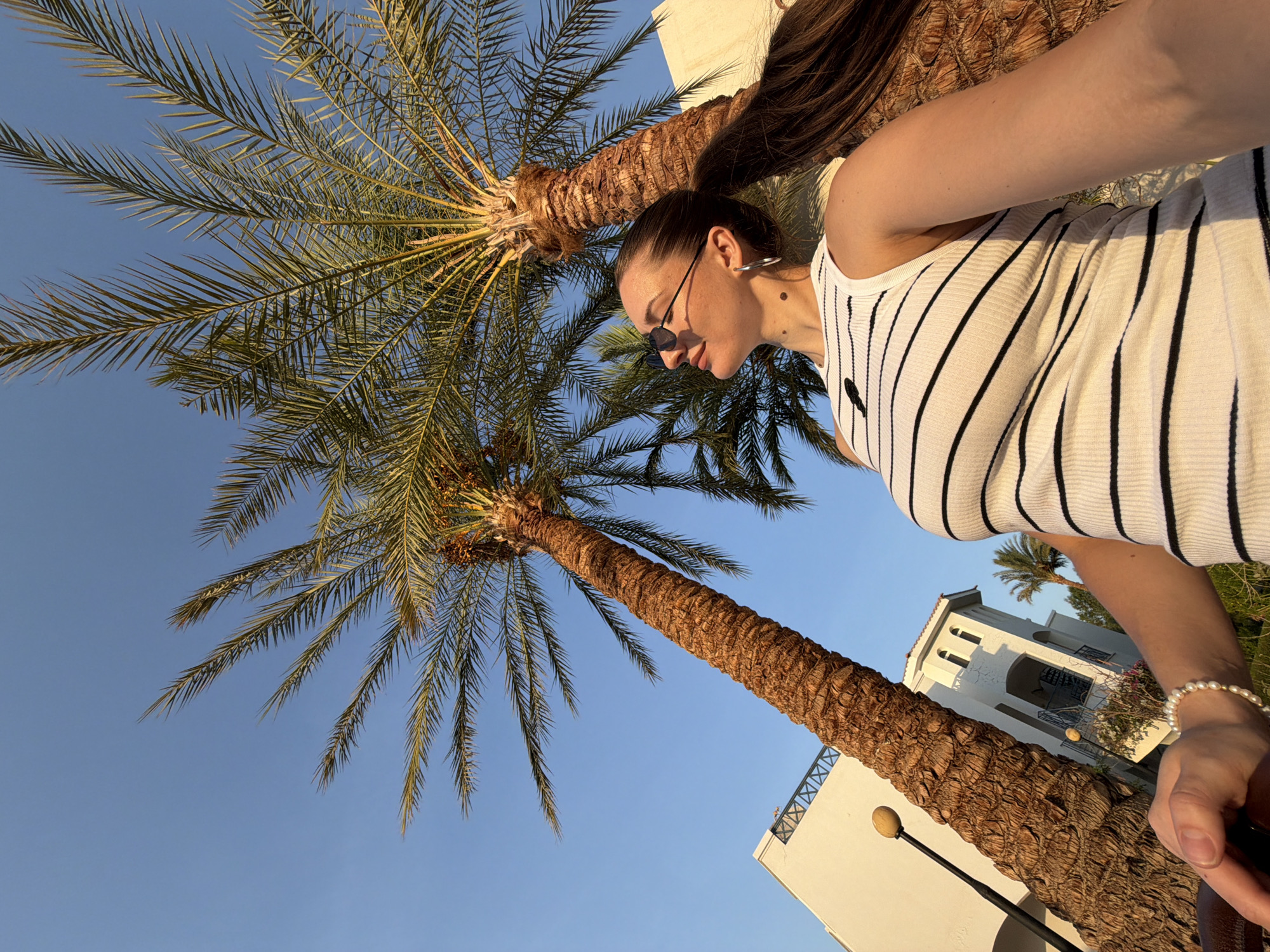 A woman stands outdoors in Sharm El Sheikh, Egypt, with two palm trees and a white building in the background. The woman is wearing a white tank top with black stripes and sunglasses. She is looking up at the sky with a slight smile. The palm trees are tall and have large fronds. The building appears to be a hotel or resort, with balconies and windows visible. The sky is clear and blue, indicating a sunny day. The image is taken from a low angle, emphasizing the height of the palm trees and the woman.