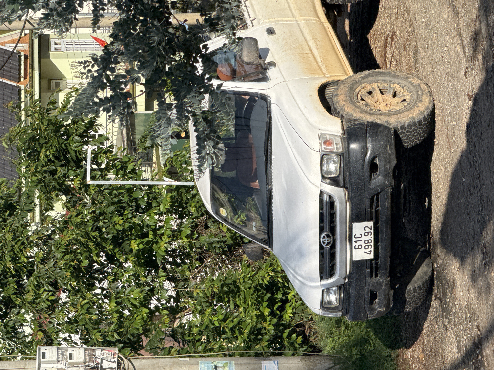 In Đa Thọ, Vietnam, a white Toyota Hilux pickup truck is parked on a gravel and dirt road, partially obscured by overgrown green foliage and a dusty eucalyptus tree. The truck is dirty, suggesting recent off-road use, and its tires are mud-caked. The license plate reads "61C 498.92". Behind the truck, a light green building with white-trimmed windows and a dark tiled roof is visible, with a small red flag on a pole projecting from one of its balconies. A utility pole with electrical meters and various wires stands to the left of the truck, adding to the urban-rural blend of the scene. The lighting suggests it is daytime, with strong shadows cast on the road. There are no people visible in the image.