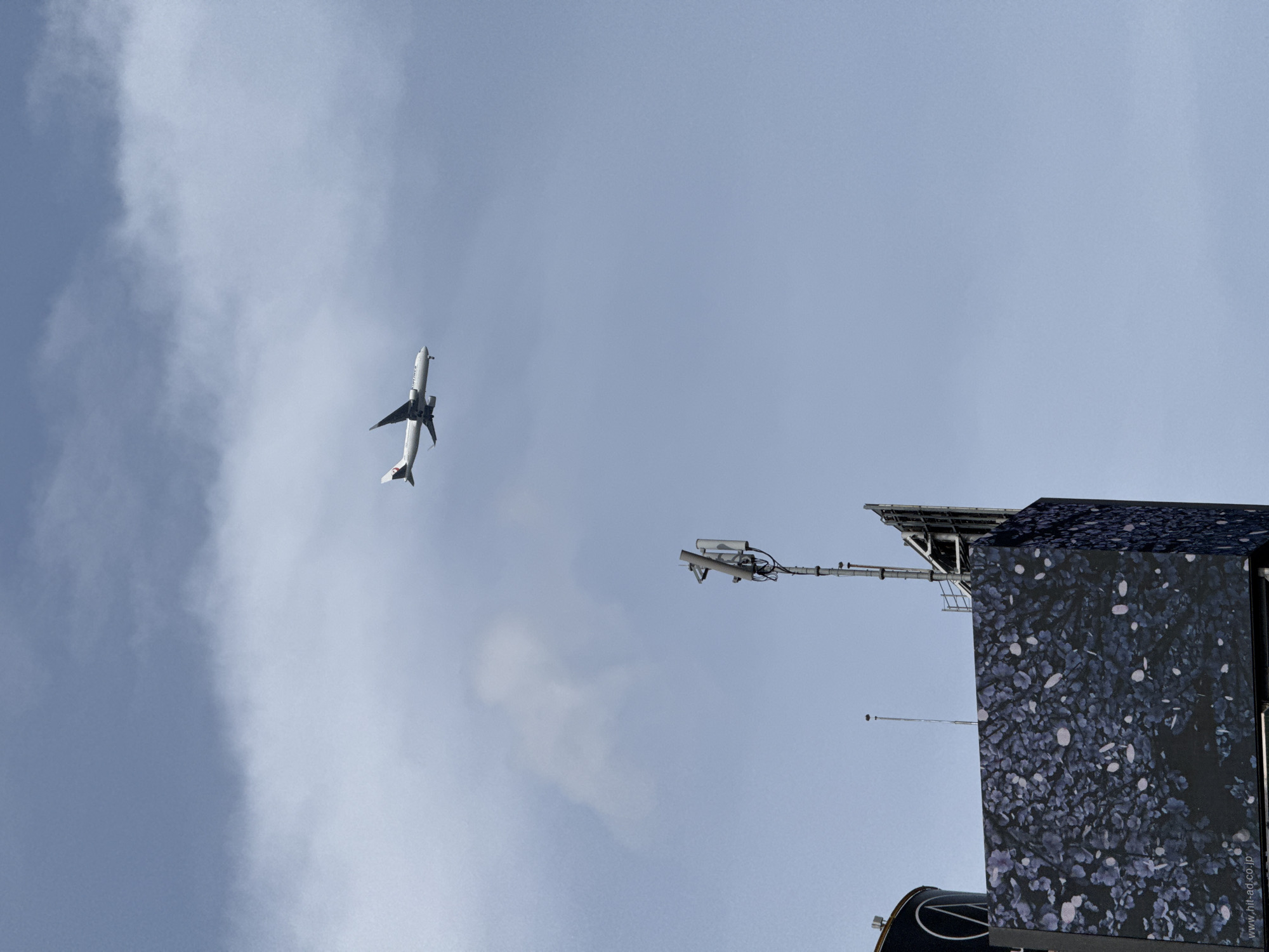 The image captures a low-angle view of an airplane flying against a backdrop of a light blue, partly cloudy sky. In the lower right corner, a section of a building or structure is visible, exhibiting a patterned exterior of leaves or flowers. A set of security cameras and mounting equipment is extending from the top of the building. The sky shows some cloud formations. It's likely Shibuya, Japan, due to the visible website address "www.nit-ad.co.jp," which is a Japanese advertisement company.