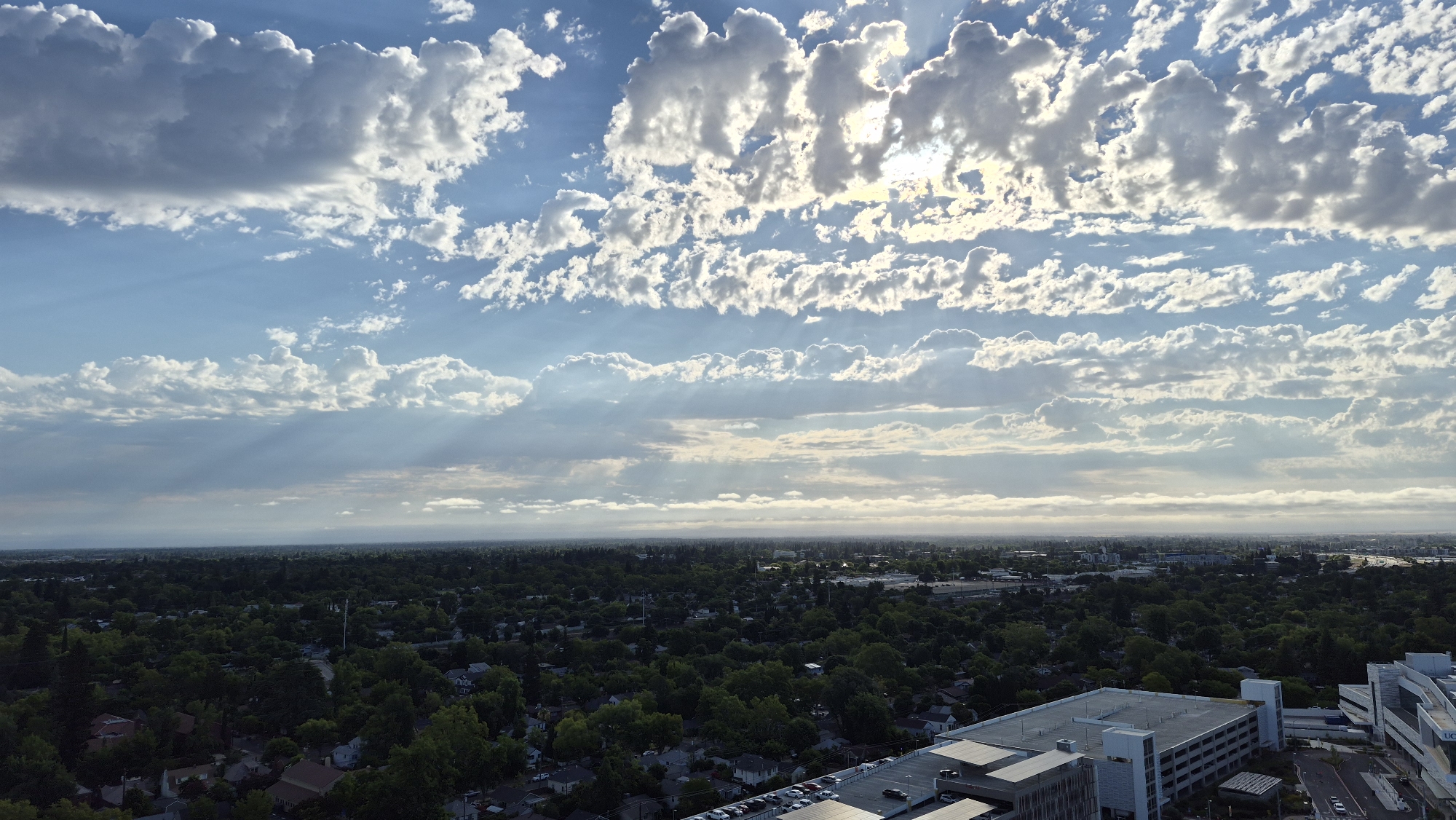 This is an aerial photograph capturing a vast landscape under a dramatic sky. The scene is dominated by a dense canopy of green trees stretching across rolling hills, interspersed with houses and buildings. In the foreground, a modern, multi-story parking garage and adjacent structures are visible, suggesting an urban or suburban setting. The sky is a striking canvas of blue, filled with an abundance of clouds. Large, fluffy white clouds are scattered throughout, while thinner, layered clouds create a textured effect across the horizon. Sunlight filters through the clouds, casting distinct rays that illuminate parts of the sky and landscape, indicating a dynamic weather pattern, possibly during sunrise or sunset given the soft, diffused light and the presence of sunbeams. The overall impression is one of natural beauty and human habitation coexisting. The location is identified as Sacramento, United States of America. There is no visible text in the image.