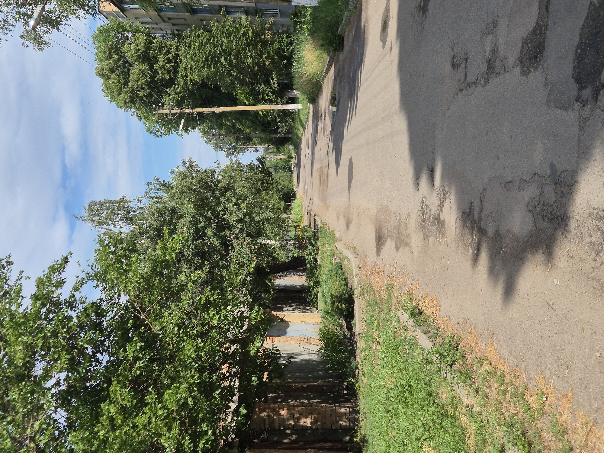 The image depicts a street scene in Sozonivka, Ukraine, under a partially cloudy blue sky. The road surface is asphalt with visible patches and puddles of water, suggesting recent rain or poor drainage. On the left side of the street, a row of brick garages with wooden or metal doors is lined with green grass and some bushes. Overhead, mature trees with dense green foliage cast shadows on the road.
On the right side of the street, tall trees line the pavement, interspersed with utility poles supporting power lines. Further down the street, a multi-story apartment building with visible windows can be seen. The overall impression is that of a residential street in a somewhat rural or suburban setting, possibly in the outskirts of a town or city, given the presence of garages and apartment buildings alongside green spaces. There are no people or vehicles visible in the frame.