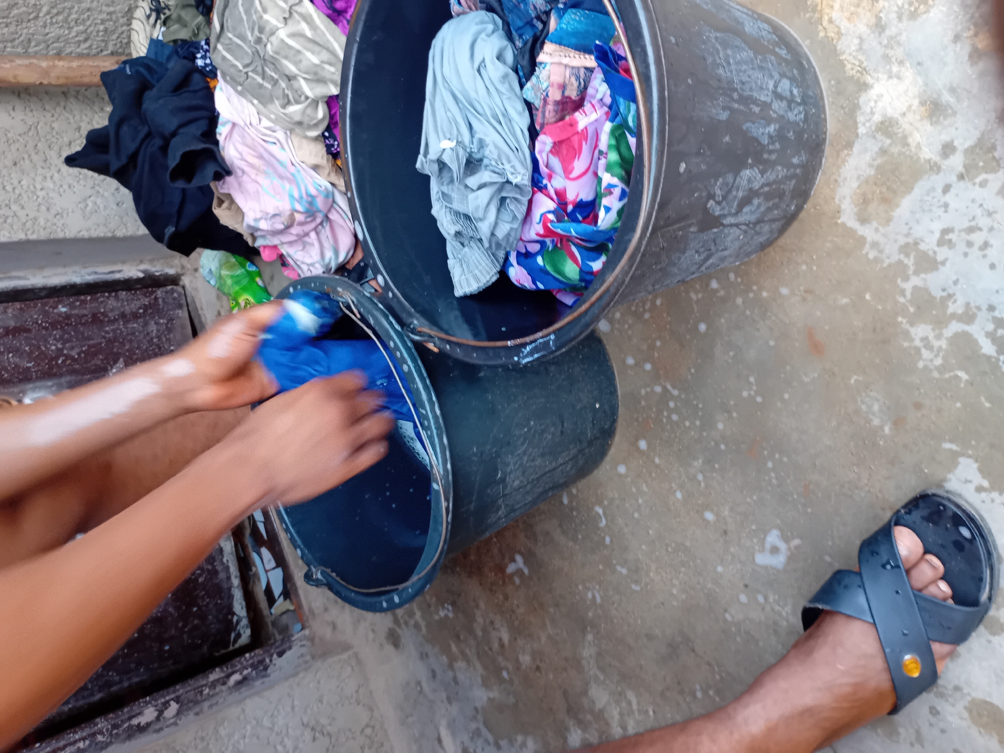 In Jauro Yinu, Nigeria, a person's hands are actively washing clothes in a large black bucket filled with soapy water. The person is wearing a black sandal on their left foot. Next to the bucket of washing, another large black bucket is filled with a pile of laundry. Various garments are visible, including a dark blue garment being held by the person, a light pink and blue striped shirt, a gray garment, and a colorful floral-patterned fabric. The ground appears to be concrete and is wet, with suds from the laundry. In the background, a portion of a wall and a wooden structure are visible, suggesting an outdoor setting. A stack of clean clothes, including dark blue, pink, and gray items, rests near the buckets.