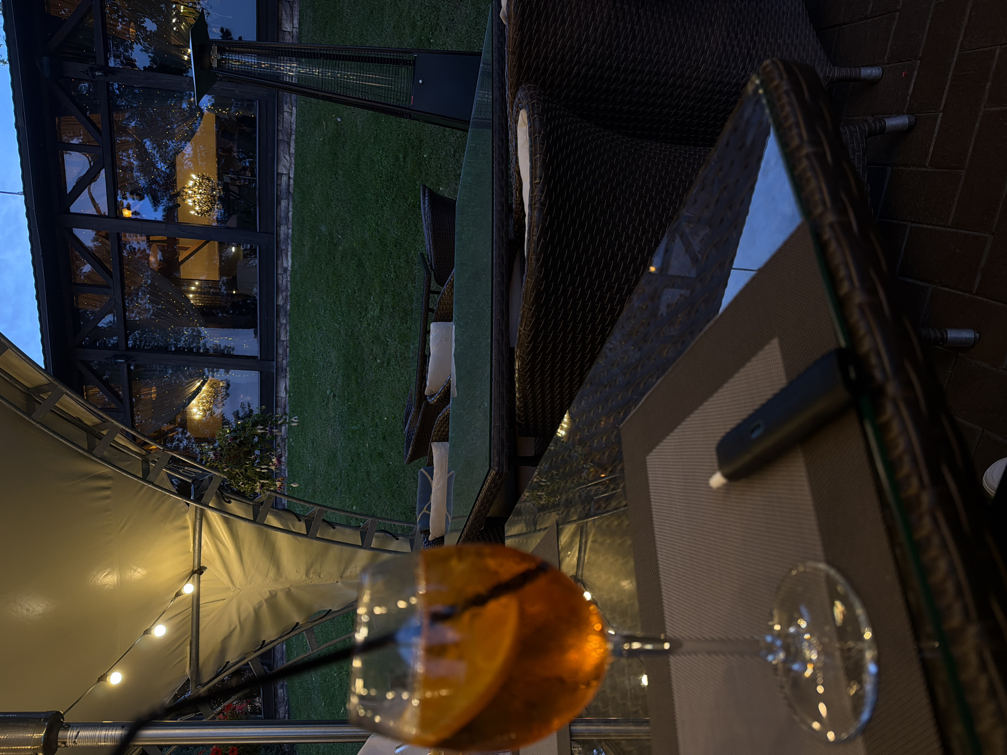 The image depicts an outdoor seating area at dusk, likely a restaurant or cafe, in Izmailovo, Russia. The scene is set on a patio with a brick floor and a grassy lawn in the background. A glass-topped table is in the foreground, with a woven chair and cushions visible. A cocktail with an orange slice sits in a stemmed glass on the table, along with what appears to be a vaping device. String lights are visible under a large, white tent-like structure, suggesting an evening ambiance. Behind the table, a modern glass-enclosed structure with interior lighting and a chandelier is visible, and beyond that, a well-maintained lawn. The lighting indicates it is twilight, with the sky transitioning from blue to a darker hue. The overall impression is one of a relaxed evening outdoor dining or lounging experience. There is no visible text in the image.