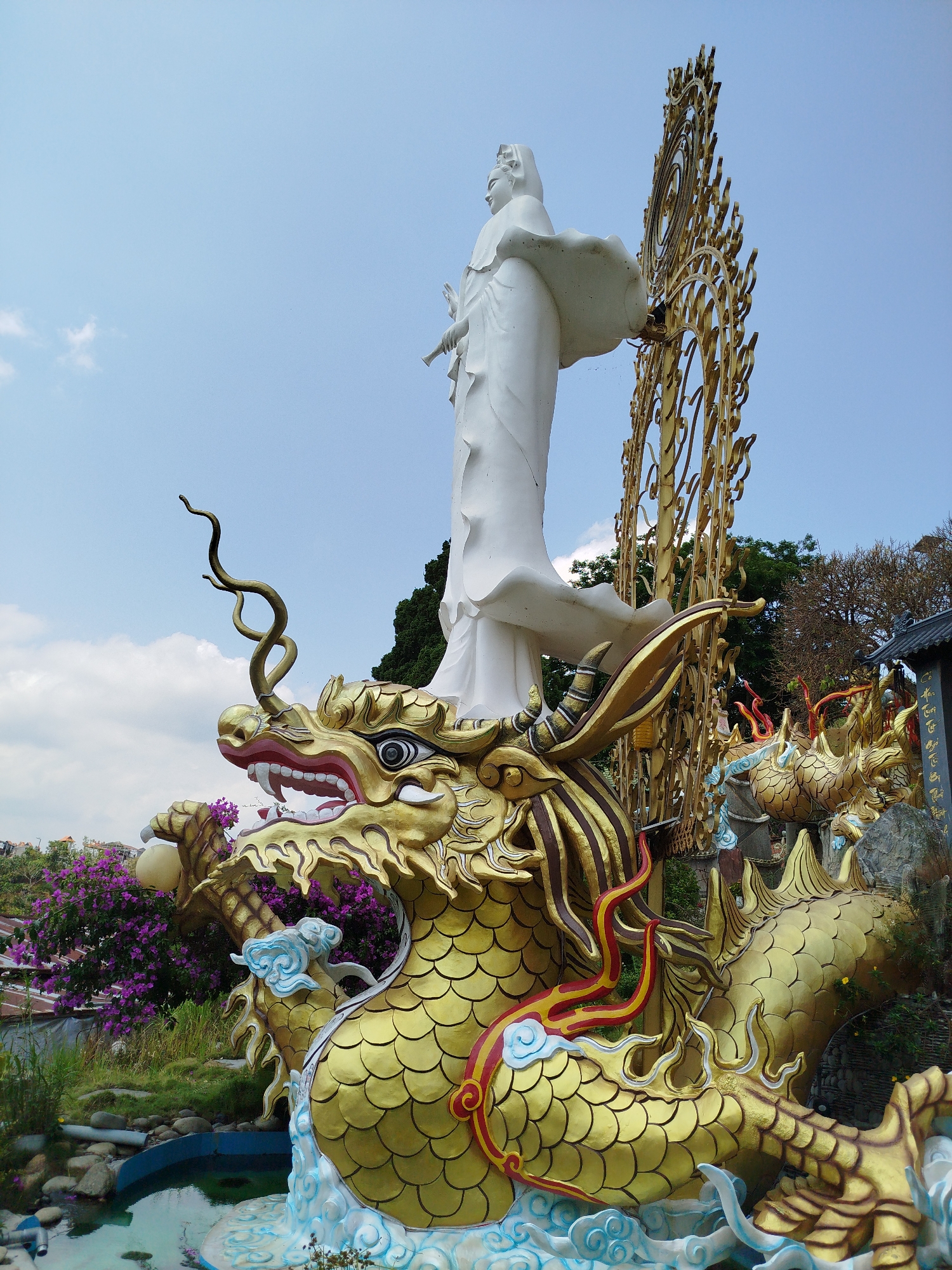The image depicts a statue of a white-robed figure standing atop a golden dragon in Diom A, Vietnam. The dragon is intricately detailed with scales, curling horns, and cloud-like accents, emerging from a small pool of water. Behind the figure is a golden, ornate halo. Purple flowering bushes surround the base of the structure. The background includes a clear blue sky with scattered clouds. Another golden dragon statue is visible behind the main dragon, along with glimpses of buildings and trees.