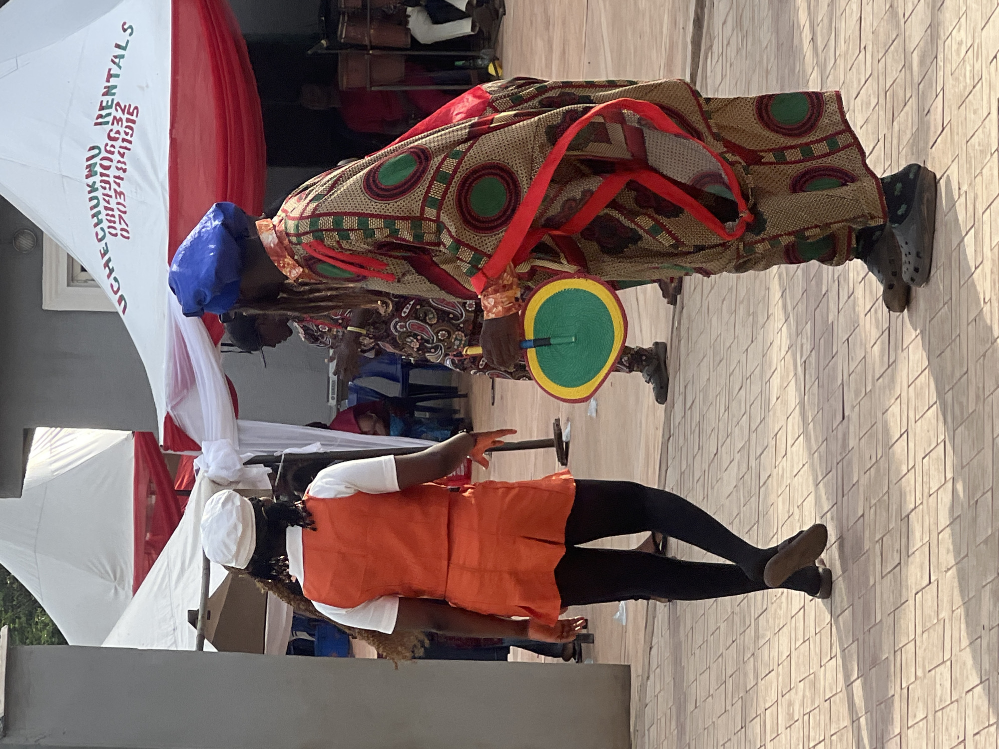 The image depicts an outdoor scene in Onitsha, Nigeria, with two individuals prominently featured. One individual, wearing a blue cap and traditional attire with geometric patterns and a red strap, holds a green and yellow fan. The other individual wears a white cap, white shirt, and an orange vest, paired with black pants. They stand on a tiled surface, which suggests a paved or constructed area. In the background, there is a white tent with red accents, with writing in black ink visible on the tent. Other figures are visible behind the first individual. The scene suggests a cultural event or celebration in an urban environment.