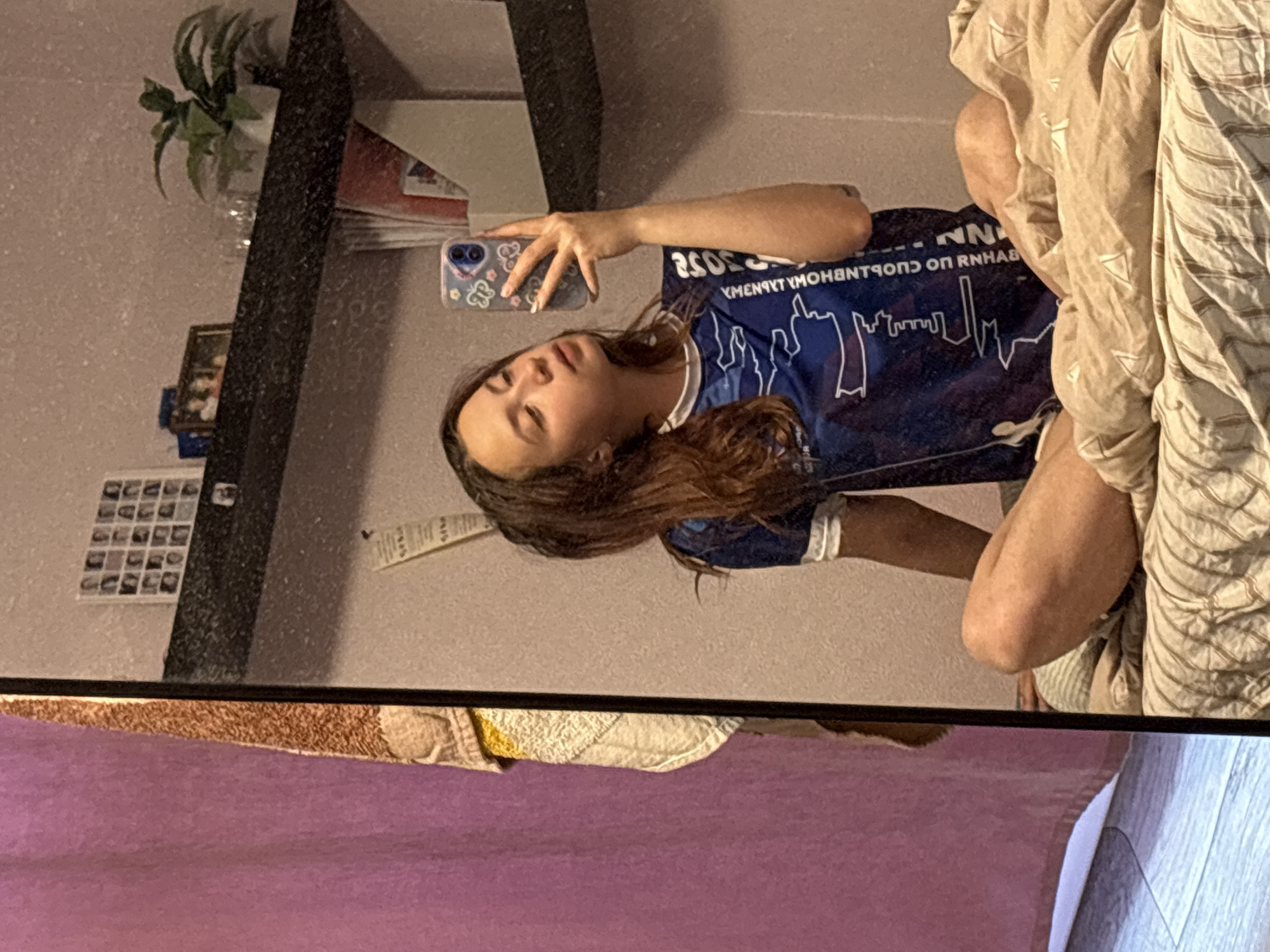 A woman is taking a mirror selfie. The woman has medium-length brown hair and is wearing a blue t-shirt with white graphics of a skyline on the front. She is holding a smartphone with a patterned case. In the background of the mirror are various items on a shelf, including a plant, books, and pictures. A collage is hanging on the wall to the left. A bed covered with bedding is to the right of the woman and in the foreground. The city of Прикамский, Russia, cannot be verified as the location from the image.
